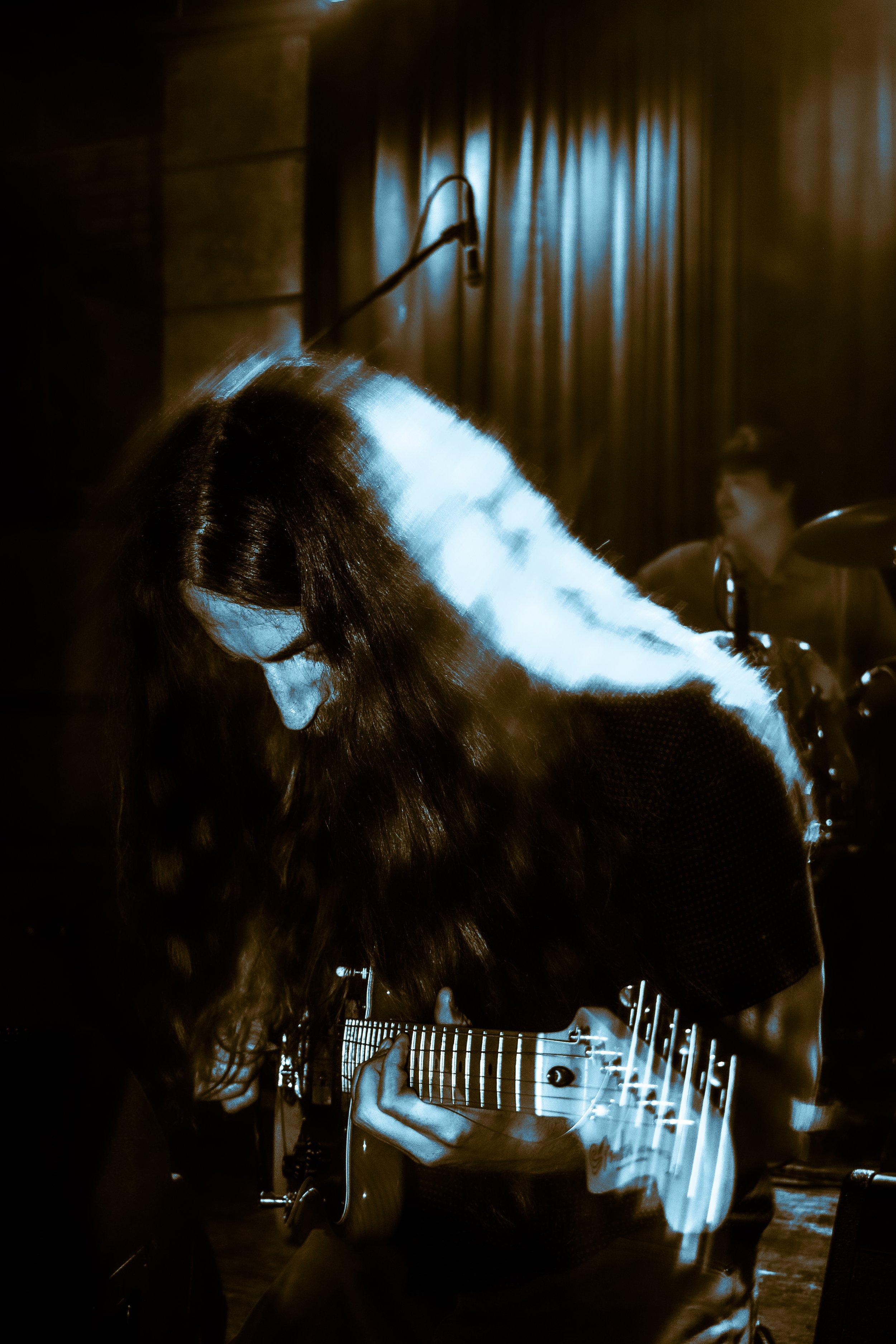 Person with long dark hair playing an electric guitar in a dimly lit room, with dramatic blue light reflections on their face and hair.