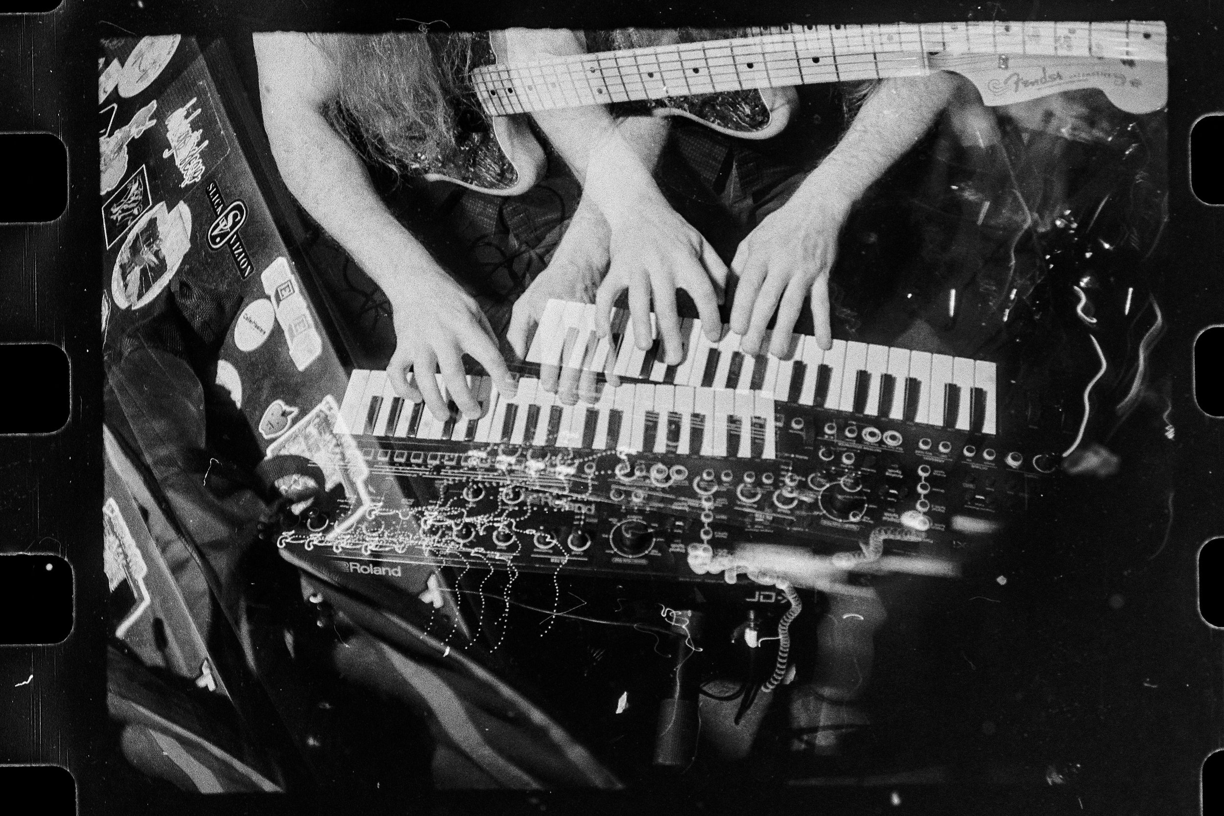 A person playing a keyboard and synthesizer with their hands. There are various electronics and stickers around, in a black-and-white photo.