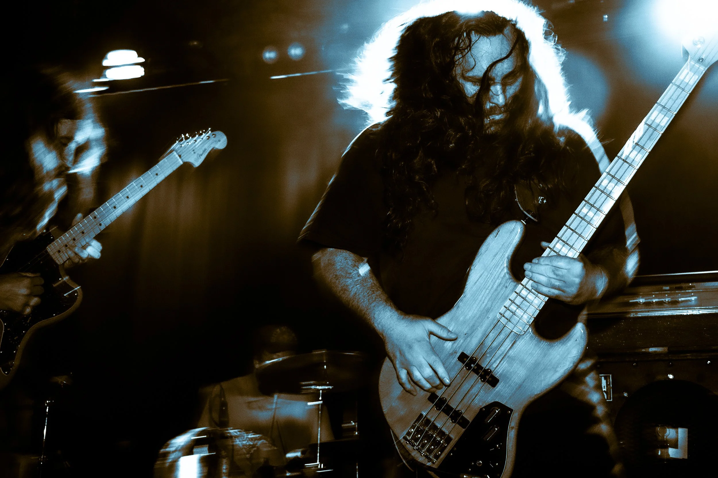 Two musicians playing guitars on stage with dynamic lighting, one with long curly hair and the other with long straight hair, in a live performance setting.