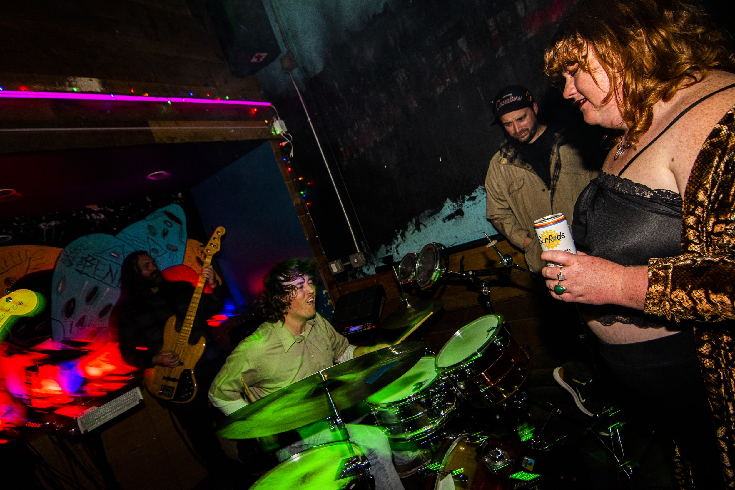 A woman with curly red hair and a leopard print jacket holding a drink and talking to a drummer, with a guitarist and another person in the background in a dimly lit, colorful bar or music venue.