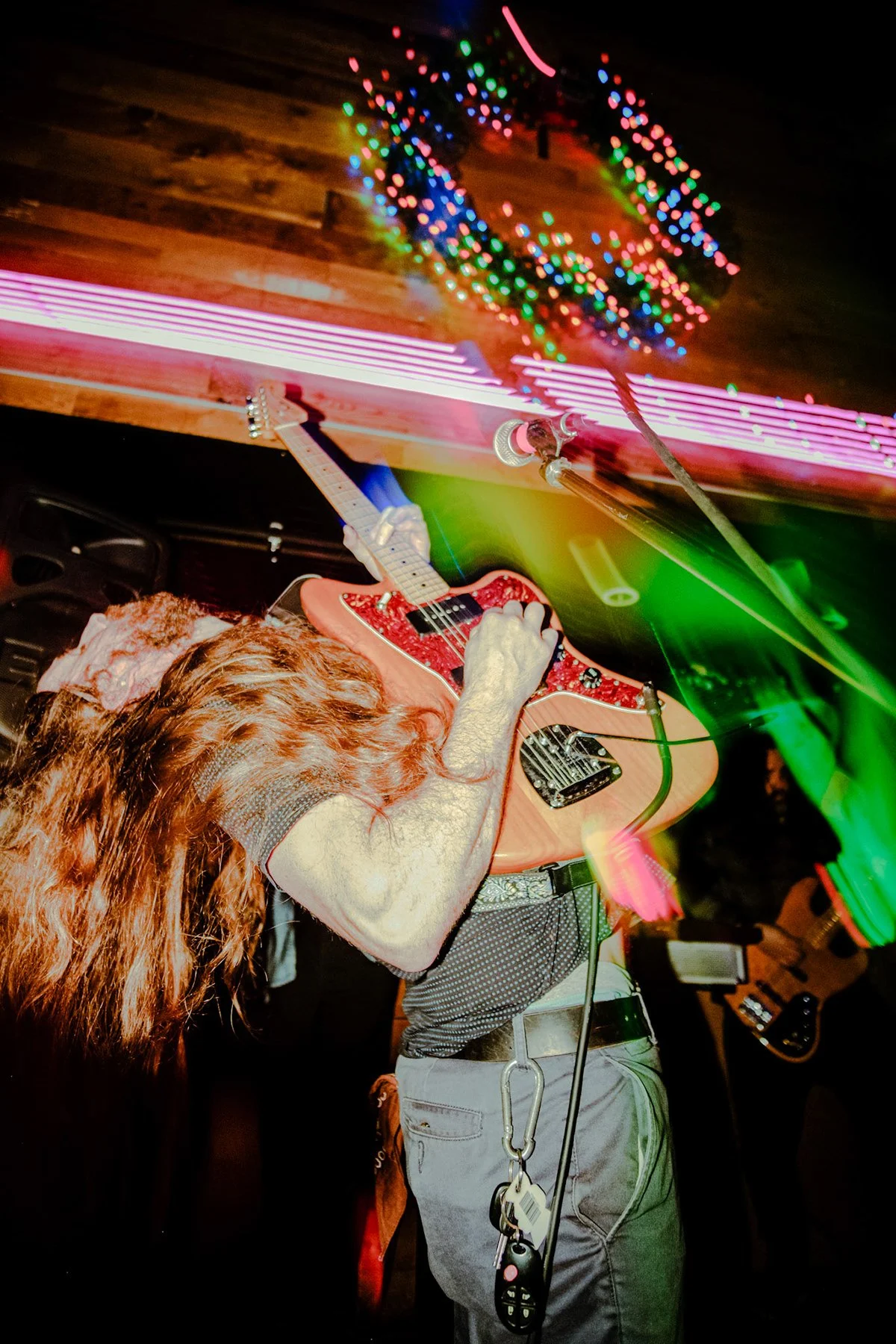 Liquid Pennies guitarist/frontman Chas Binns playing an electric guitar during a live performance with colorful lights and a wooden wall in the background.