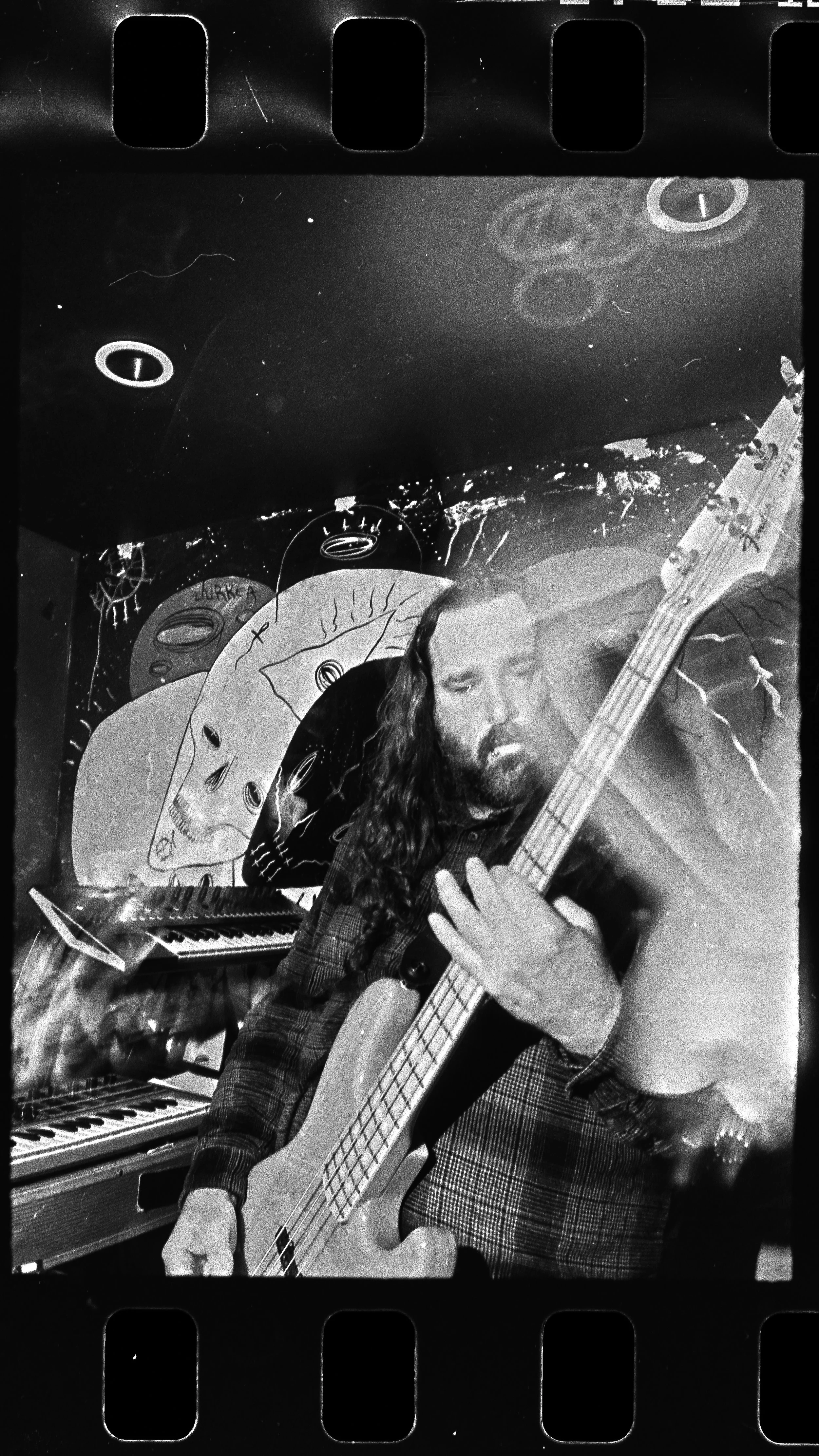 A man with long hair and a beard playing an electric bass guitar in a room with artistic and cosmic-themed wall art, including celestial bodies and abstract faces, all in black and white.