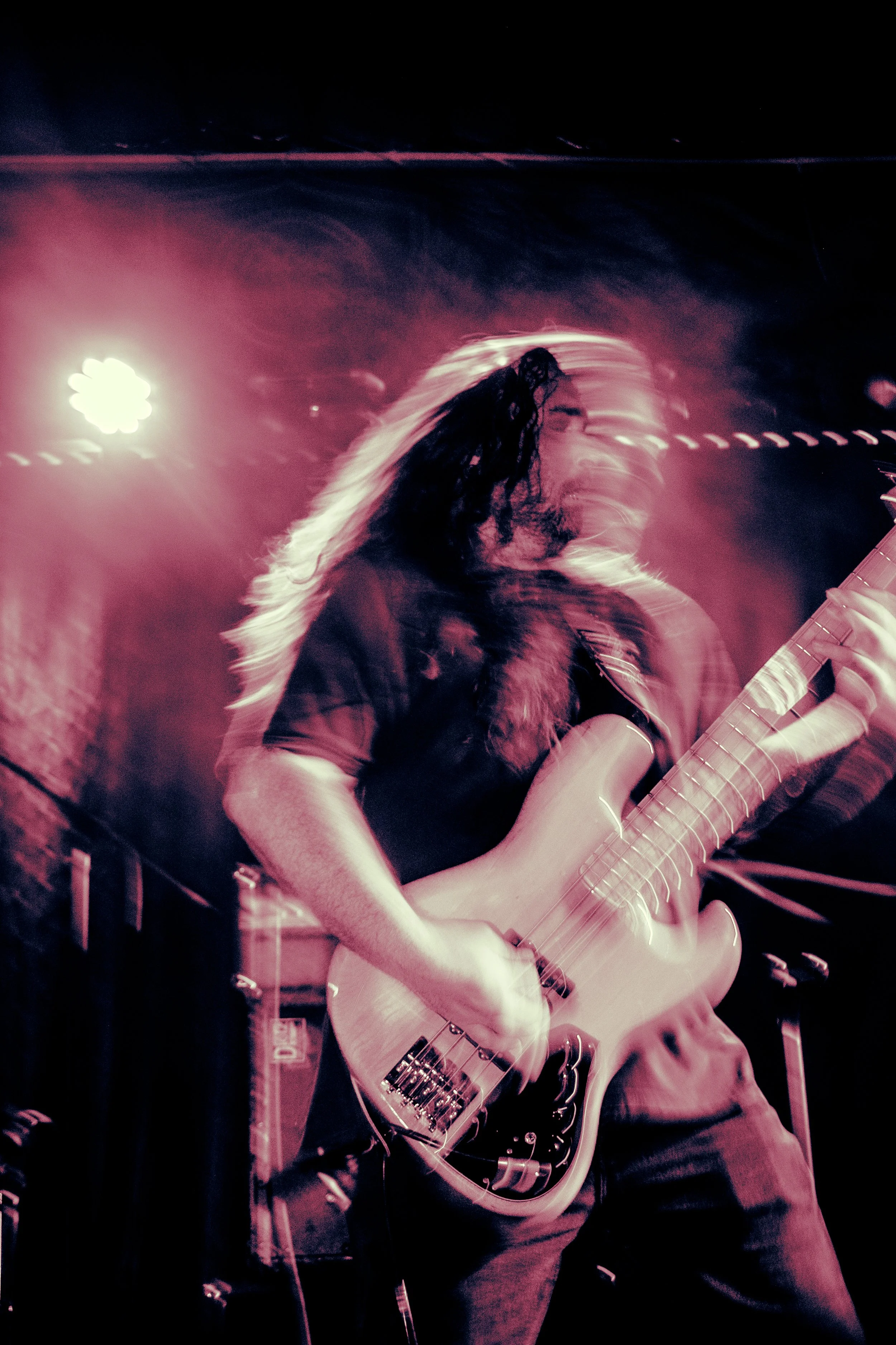 Musician playing electric bass guitar on stage with pink lighting, long hair, and a blurred motion effect.