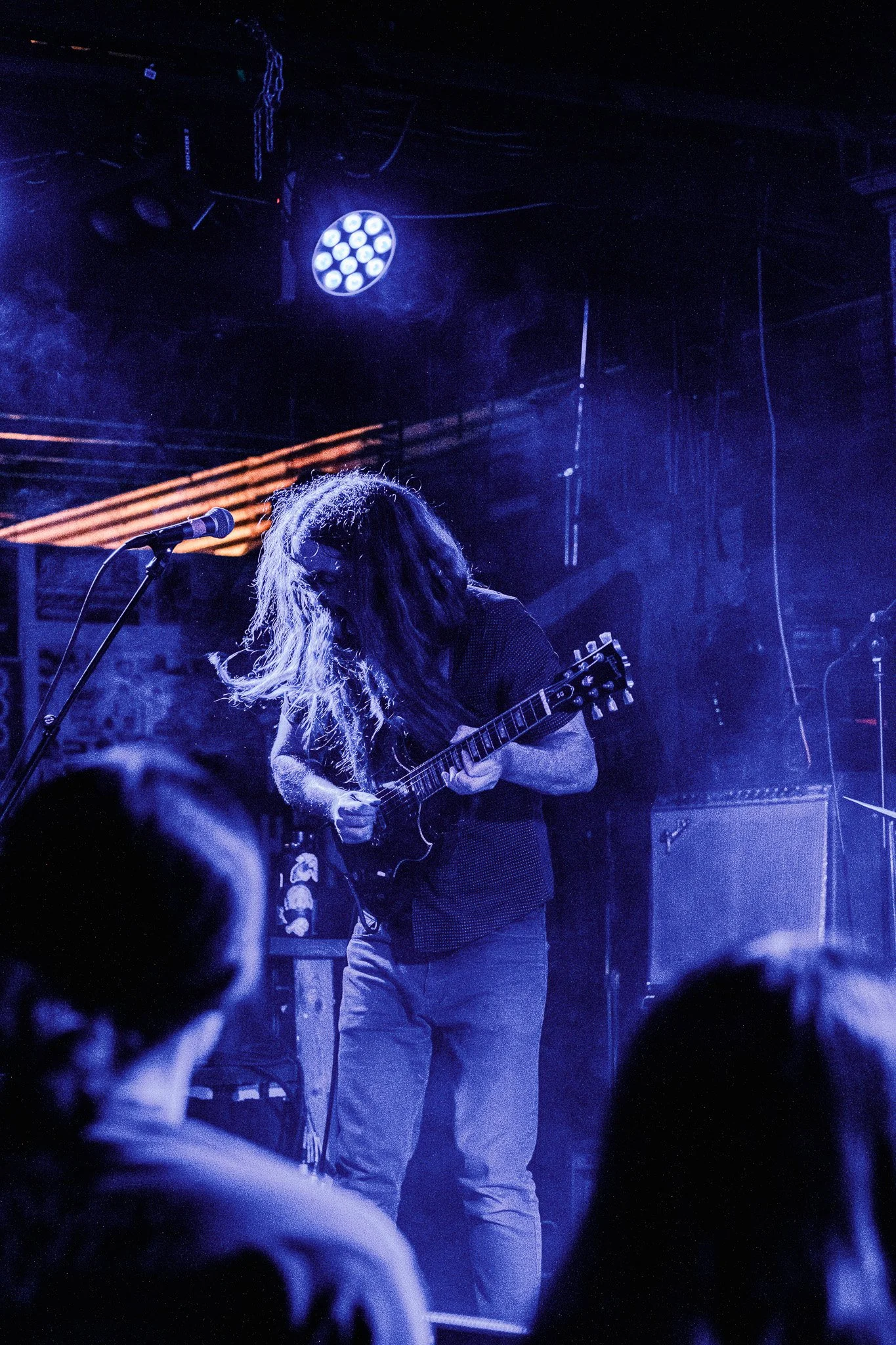 A musician with long hair playing an electric guitar on stage at a concert, illuminated by blue lights.