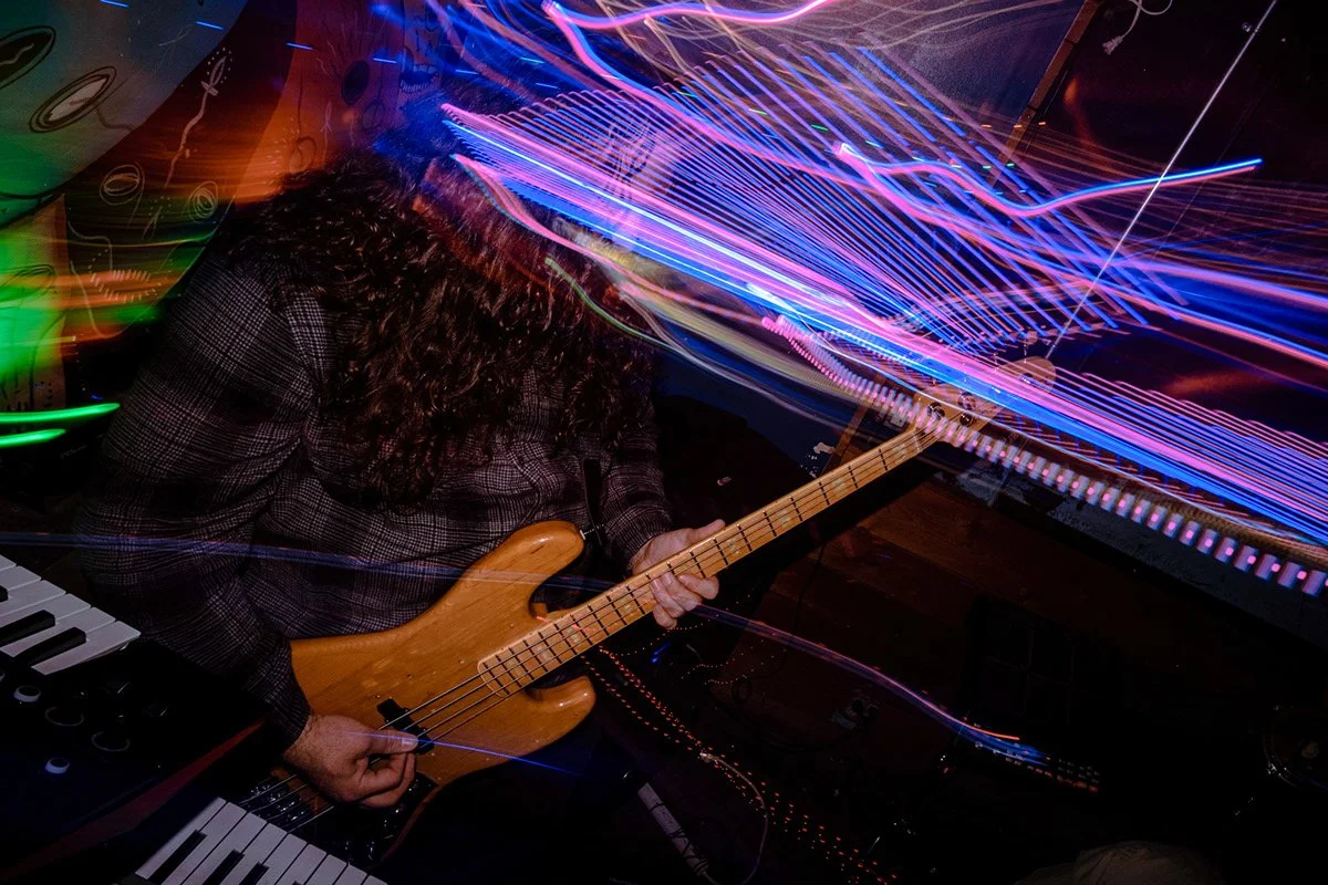 A person playing an electric bass guitar surrounded by colorful light streaks in a dimly lit setting.