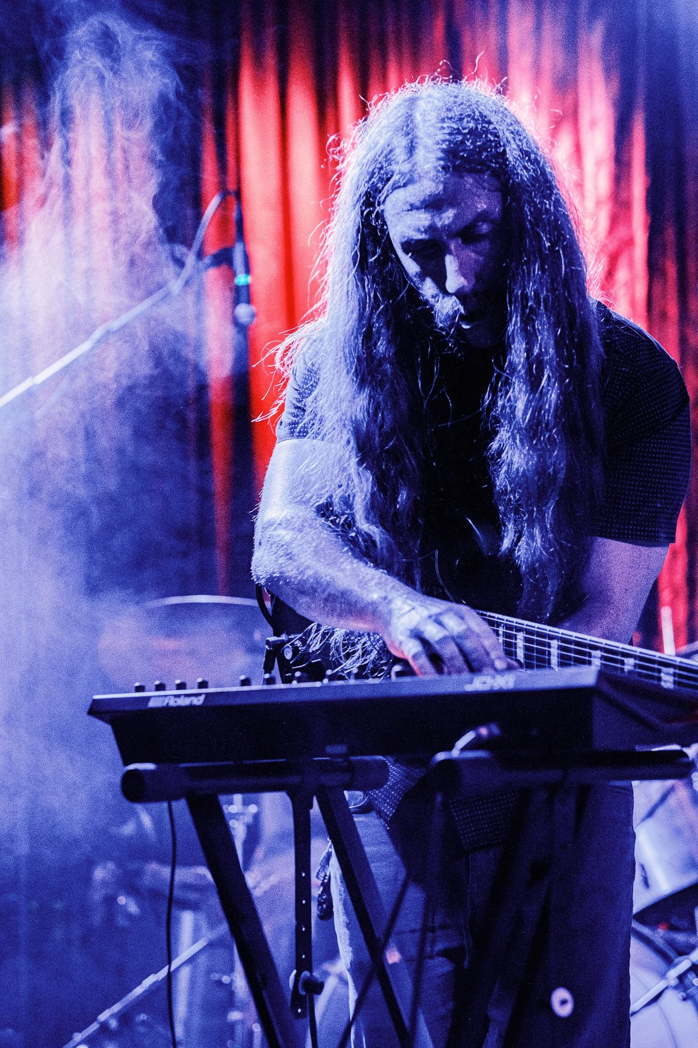 Musician with long hair playing a keyboard on stage with red curtains and purple lighting.