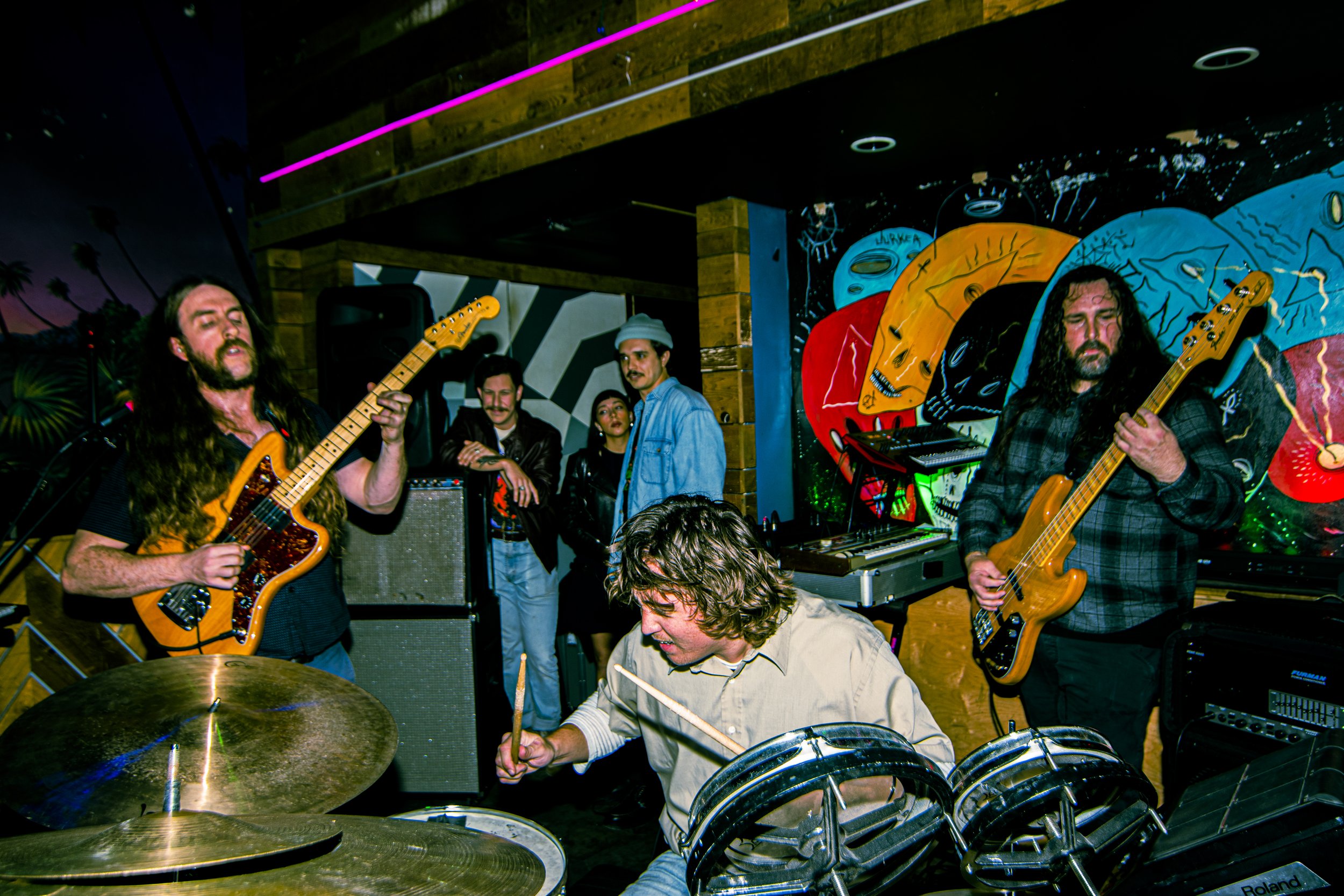 A band performing live includes two guitarists and a drummer, with a vibrant, colorful mural in the background.