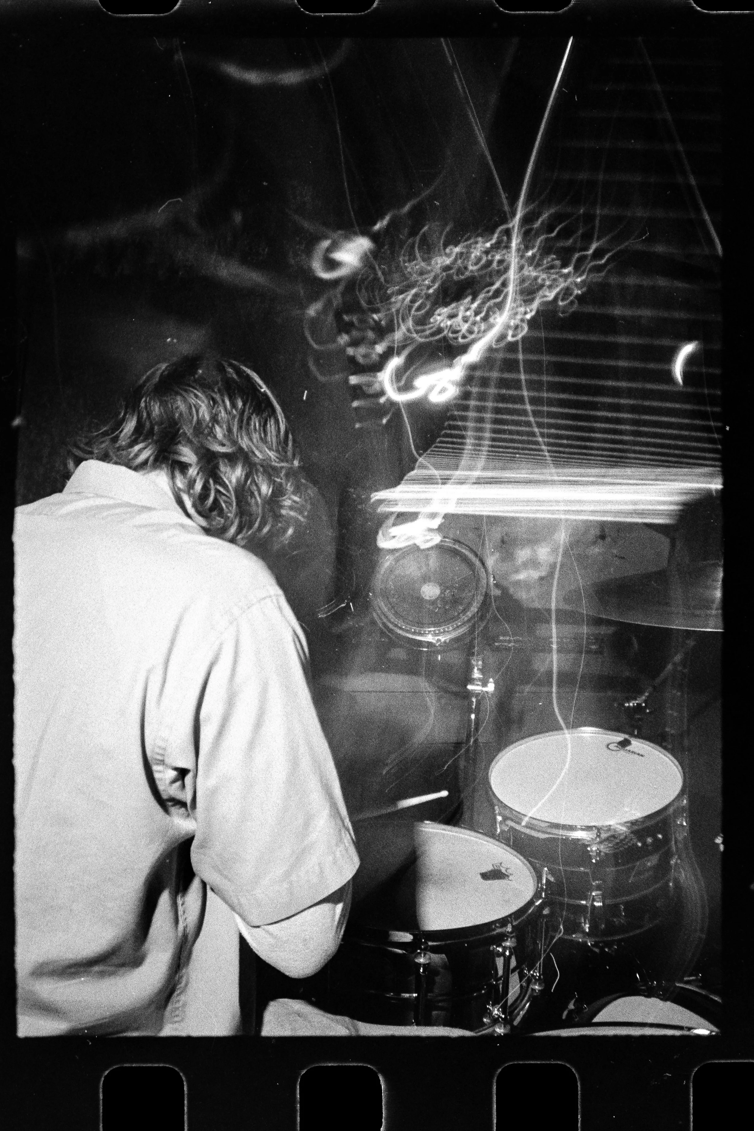 Pierson Whicker, drummer of the experimental rock band Liquid Pennies  sitting in front of a drum set, with abstract light trails across the photograph, in black and white.
