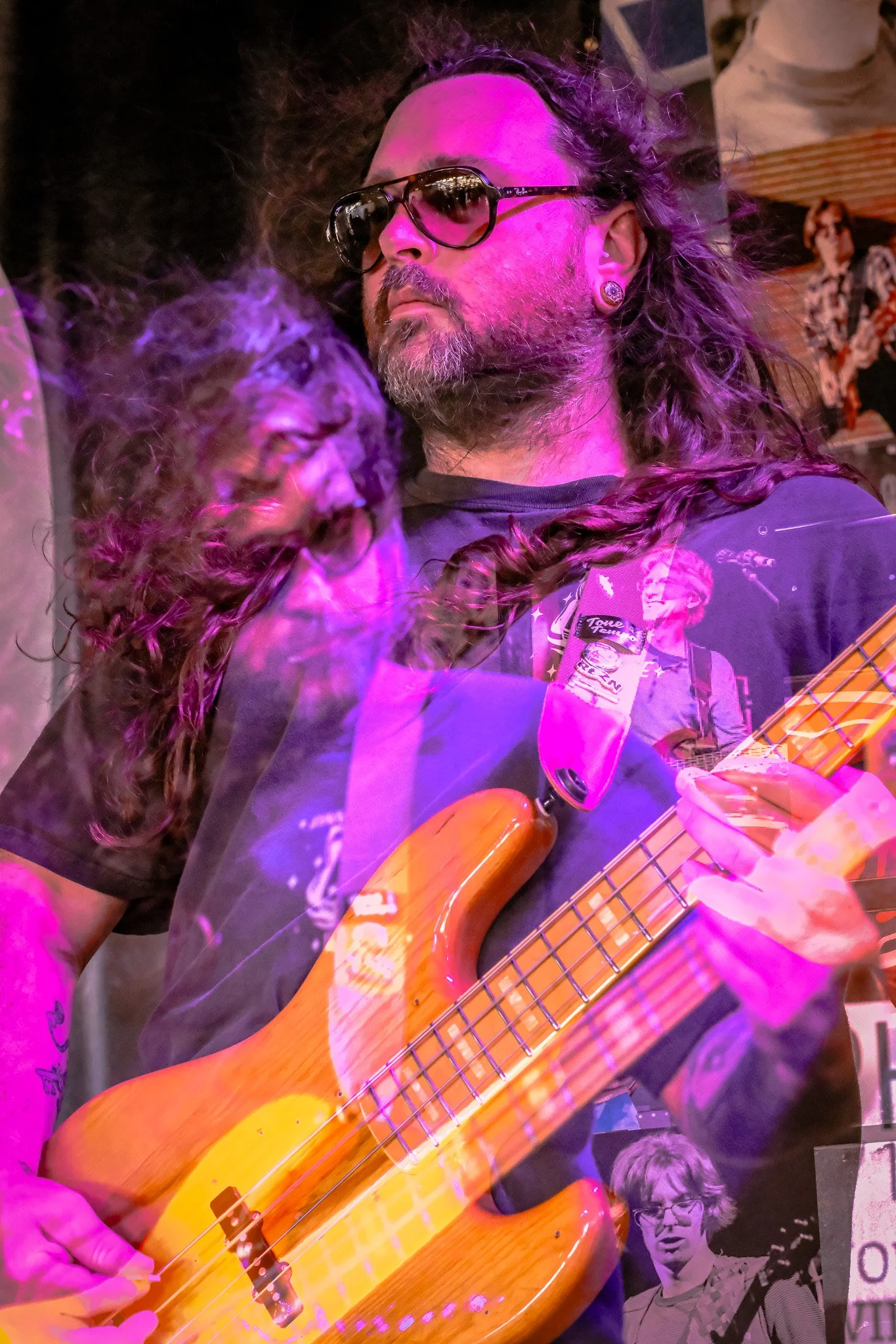 A man with long hair, sunglasses, and a beard playing a bass guitar surrounded by pink and purple stage lighting.