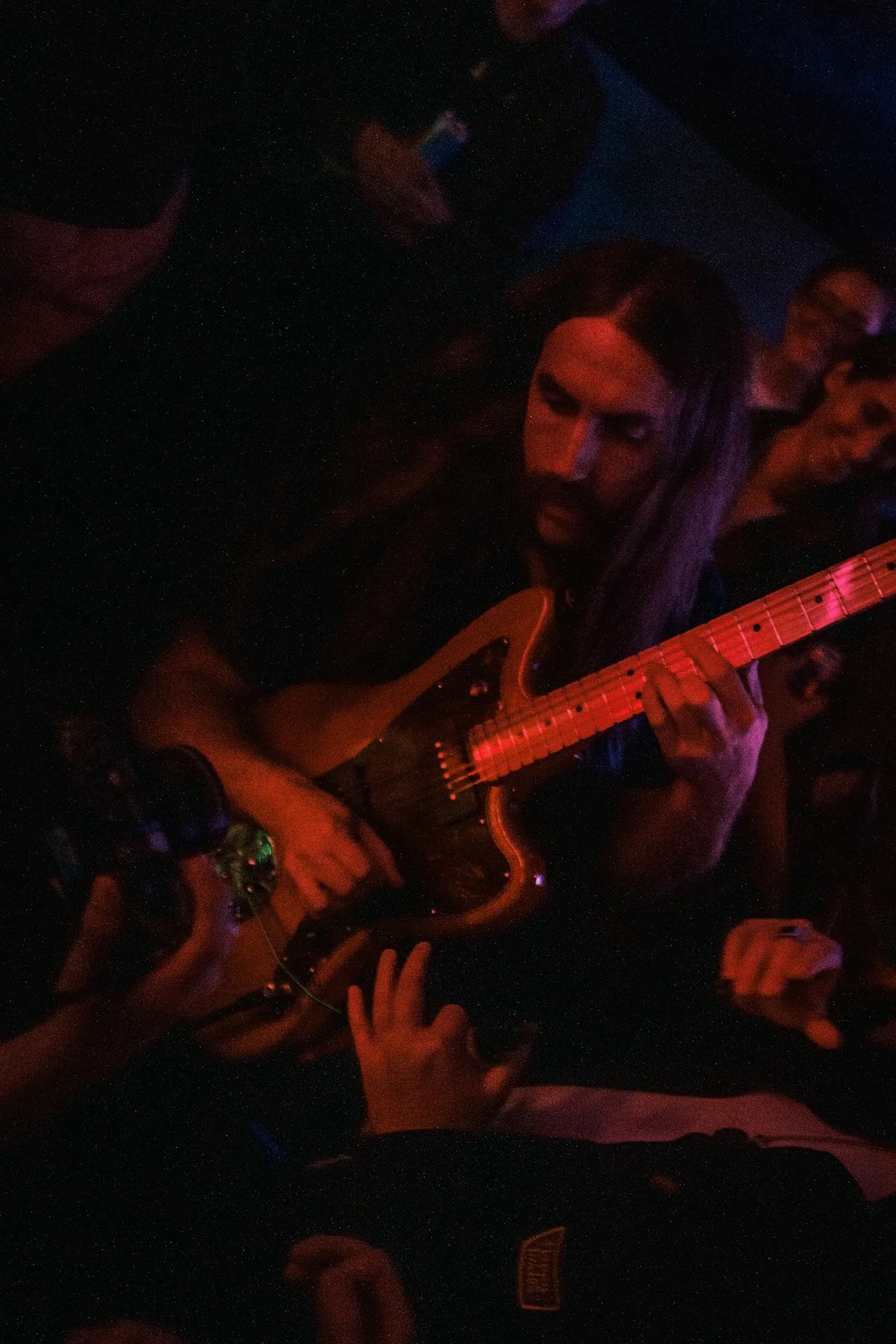 Person playing electric guitar in a dark, colorful setting with people around.