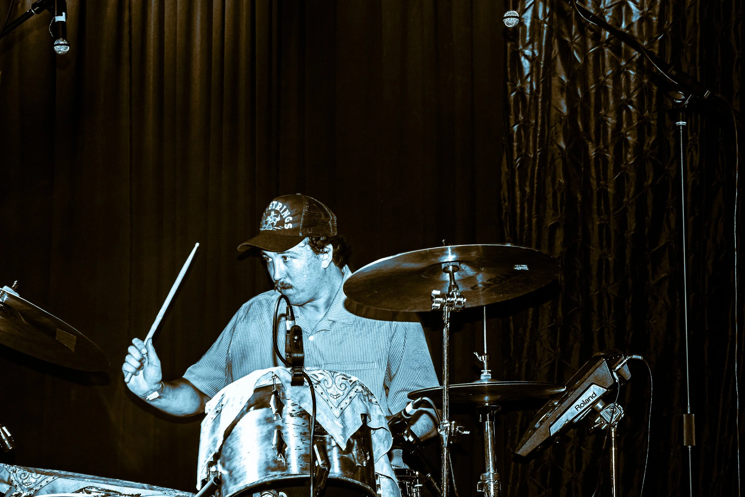 Psych rock band Liquid Pennies drummer PIerson Whicker playing drums on stage, wearing a cap, shirt, and headset, with dark curtains in the background.