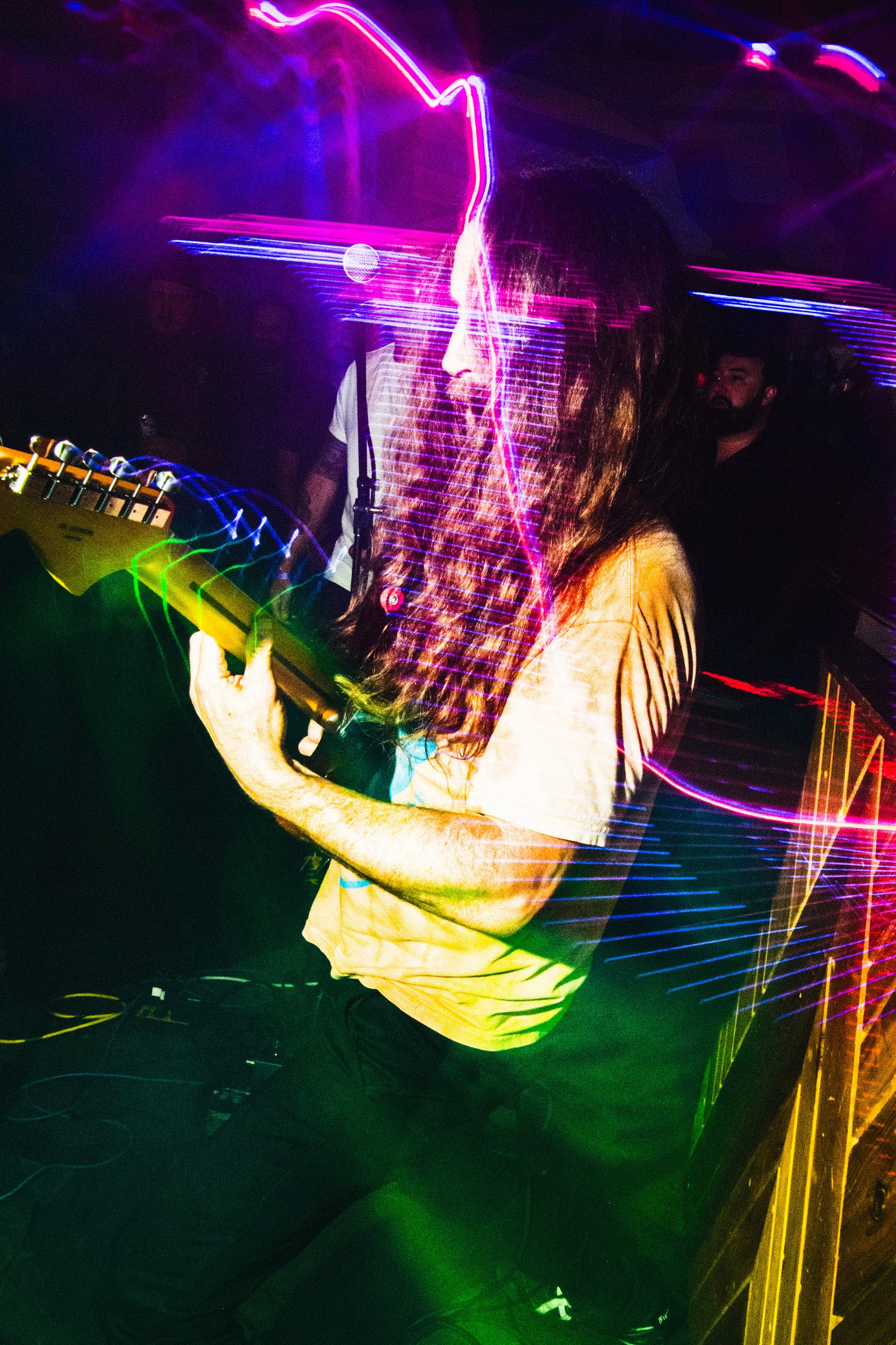 A person with long hair playing a guitar in a dimly lit nightclub with colorful neon lights.