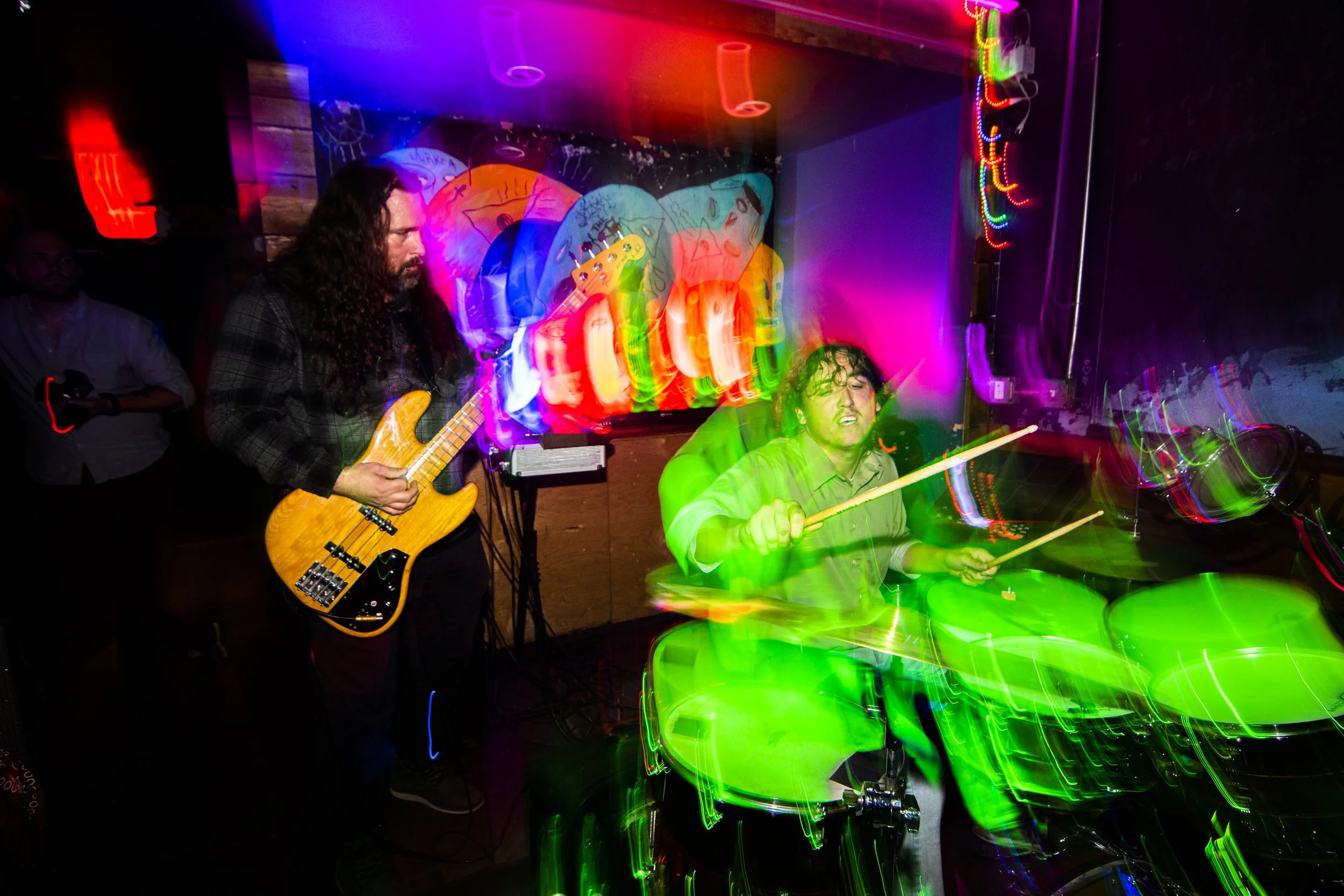 Two musicians performing with colorful light trails and a neon sign in the background.