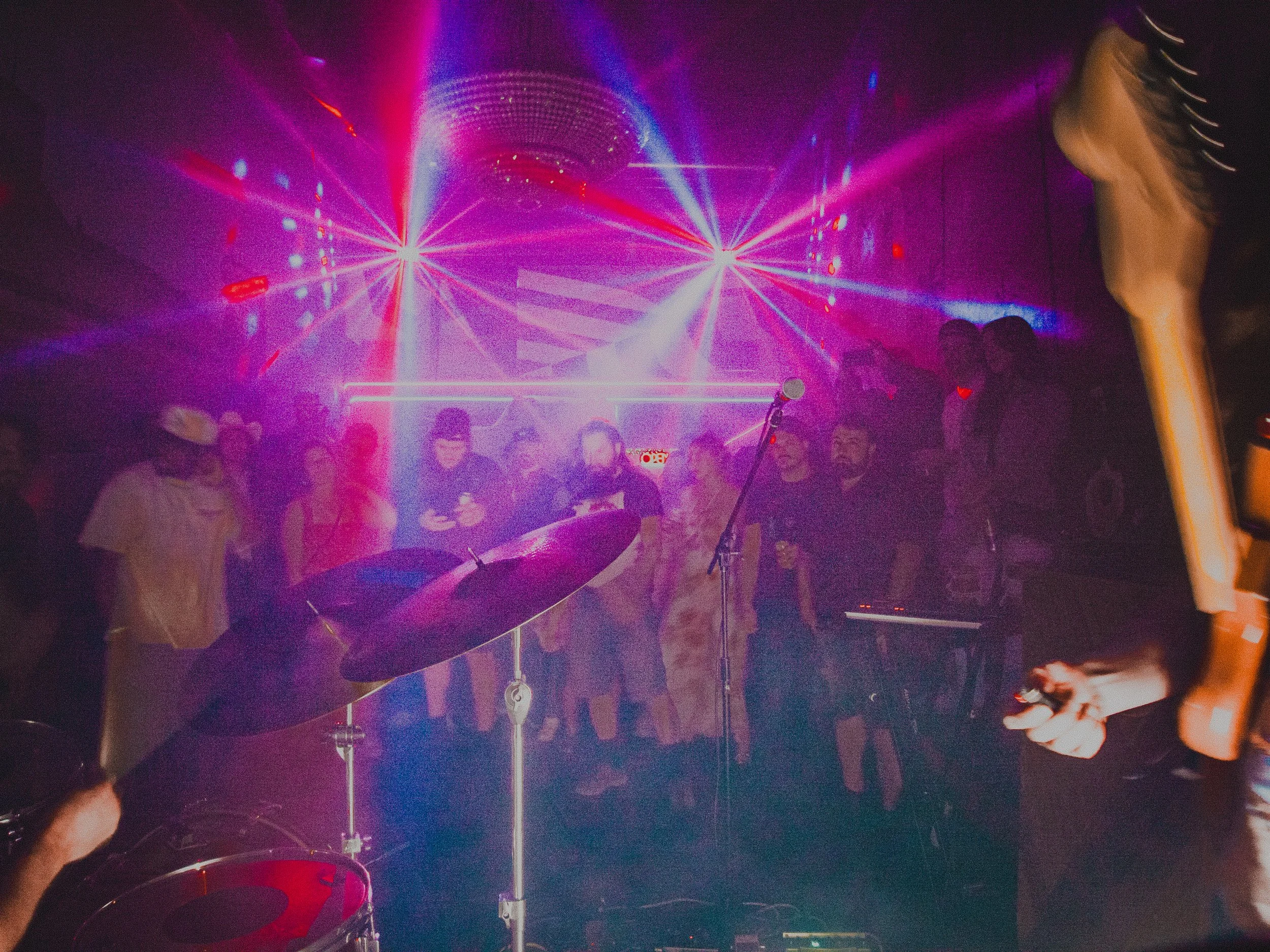 A group of people dancing and enjoying live music at a nightclub with colorful laser lights and a disco ball overhead.