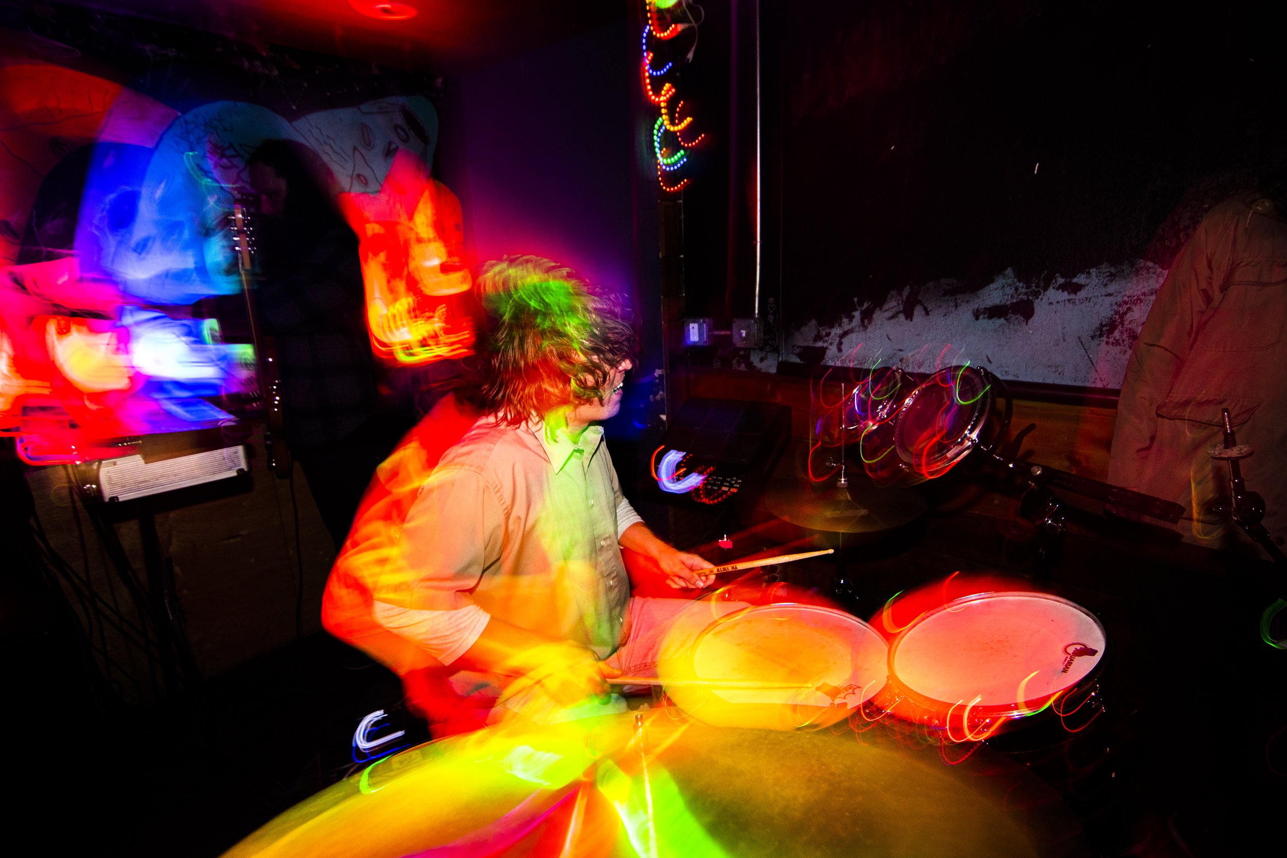 Person playing drums in a dimly lit room with colorful, blurred lights and motion effects.