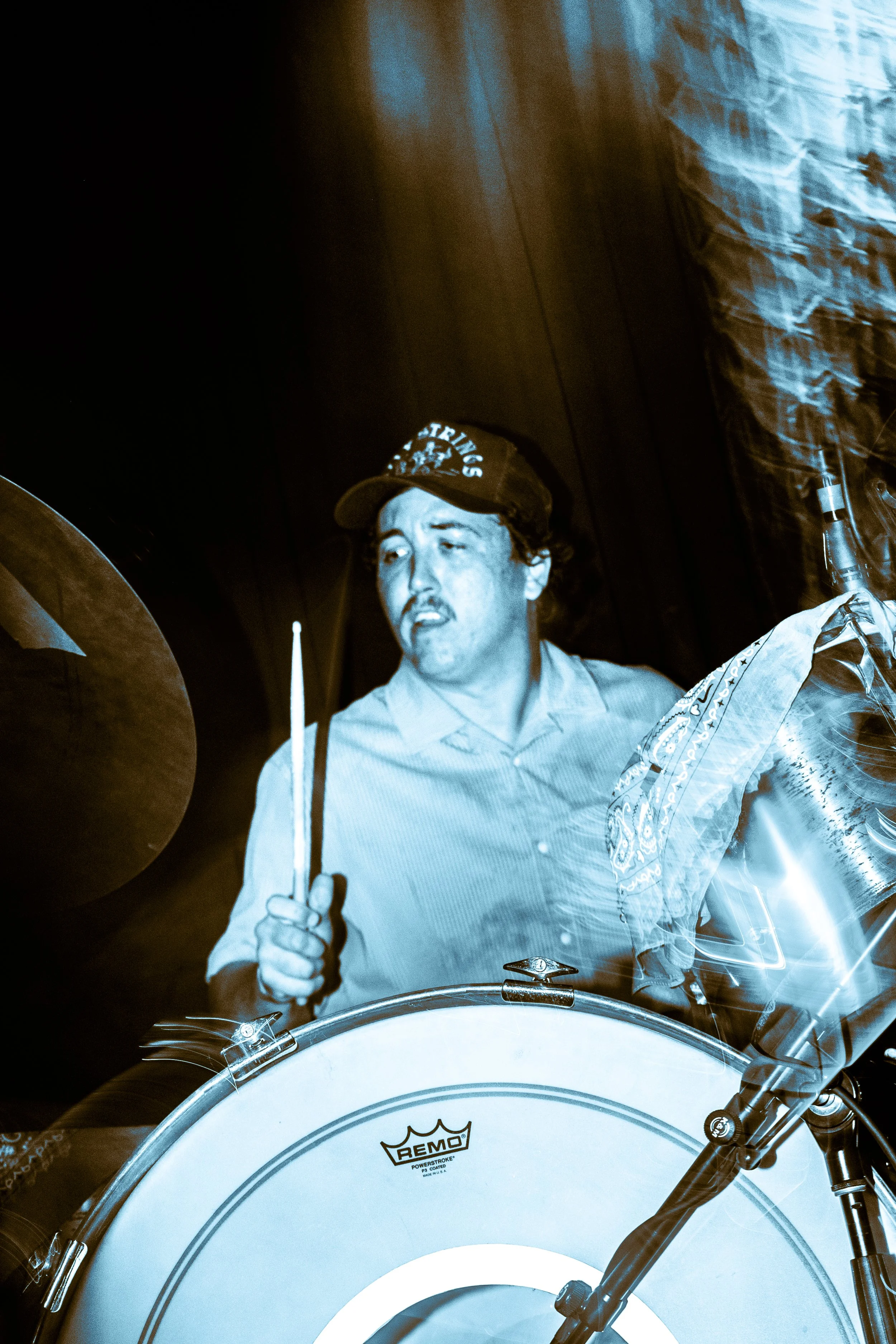 A man playing drums with a focused expression, wearing a baseball cap and a bandana around his neck, in a performance setting with stage lighting.