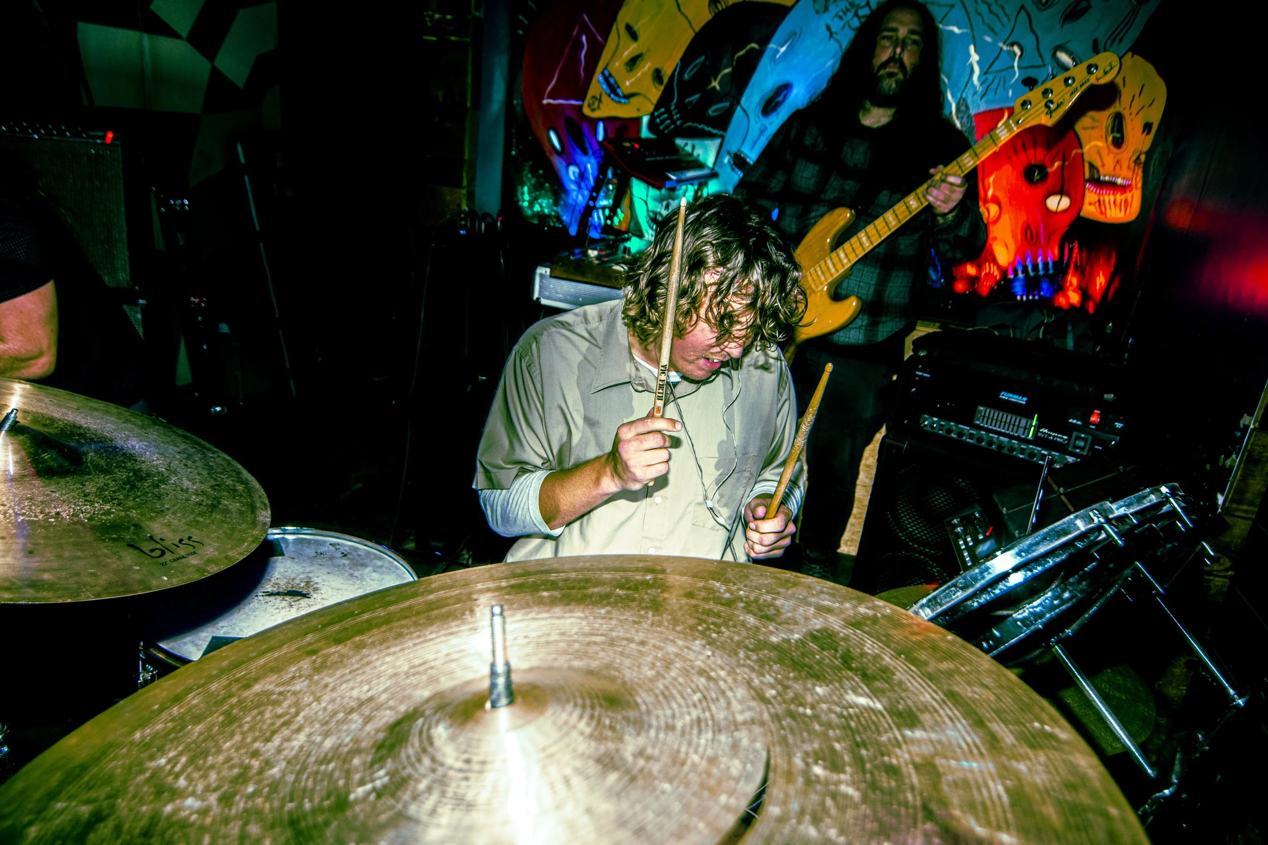 A drummer with curly hair playing drums, with a guitarist standing in front of colorful abstract art on the wall in the background. The drum set includes a noticeable cymbal in the foreground.