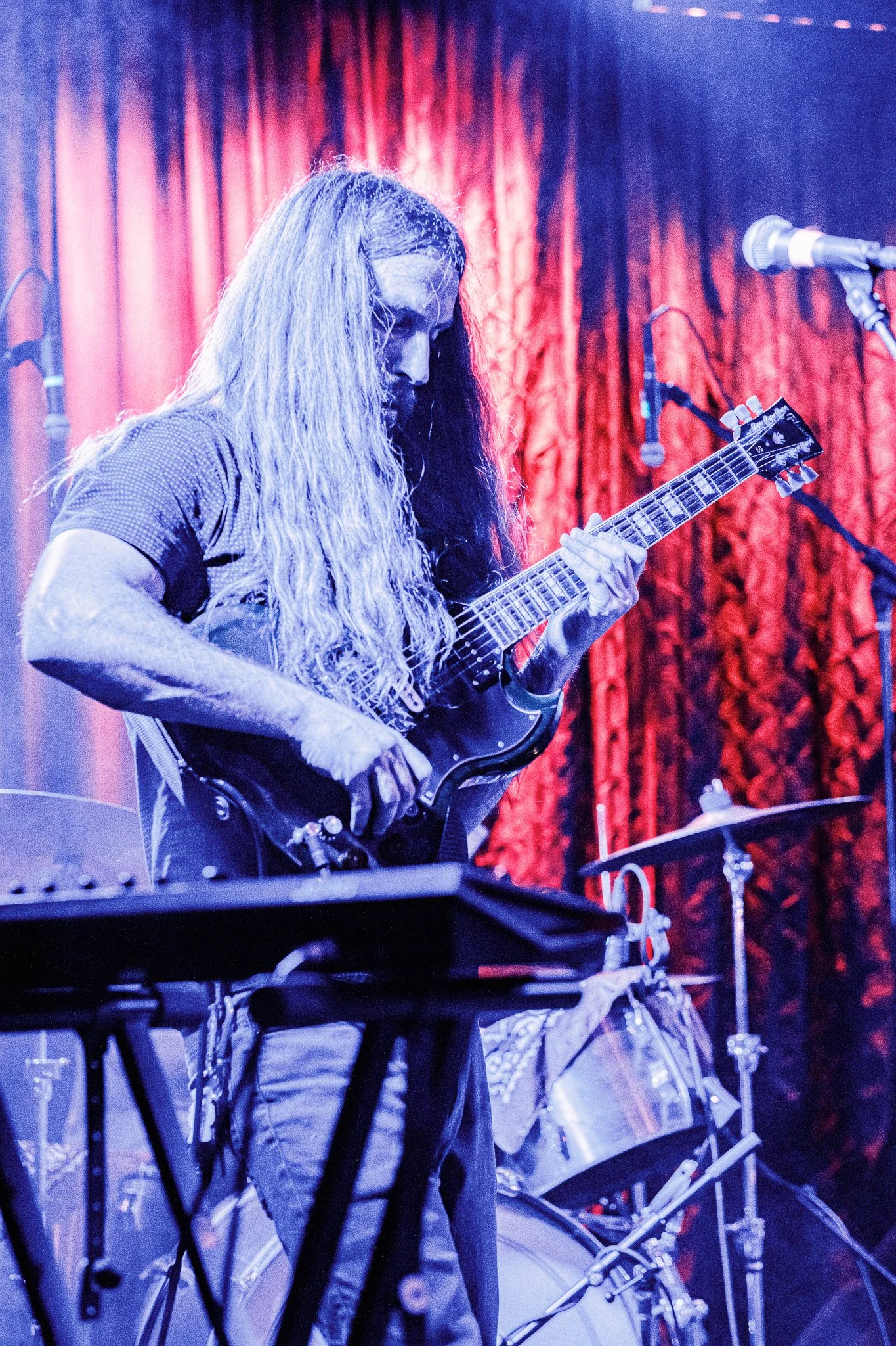 A musician with long, wavy hair playing an electric guitar on stage with red curtains in the background.