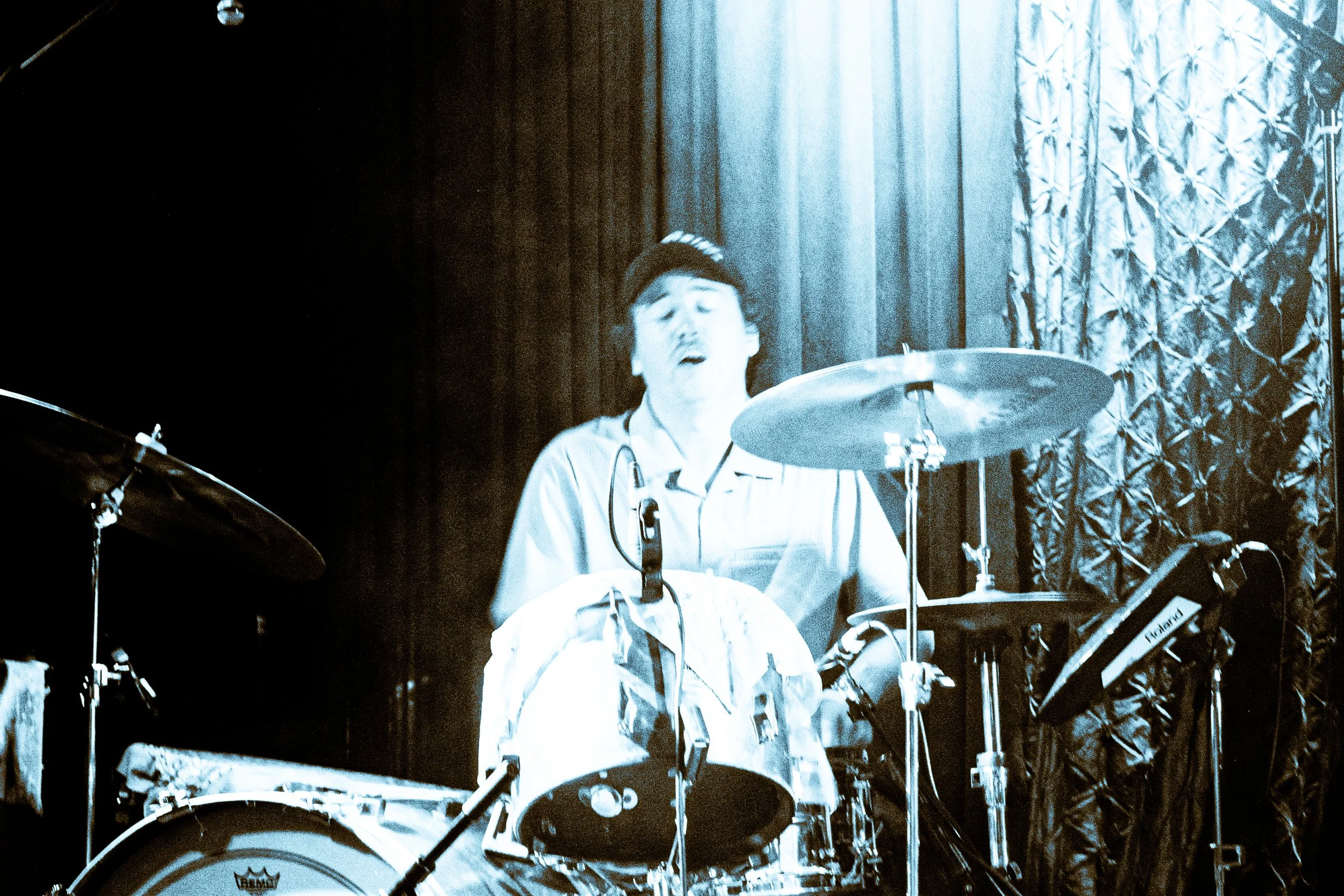 Experimental rock band Liquid Pennies drummer PIerson Whicker  playing a drum set, singing into a microphone, with a dark curtain and patterned curtain behind him, lit with a blue hue.