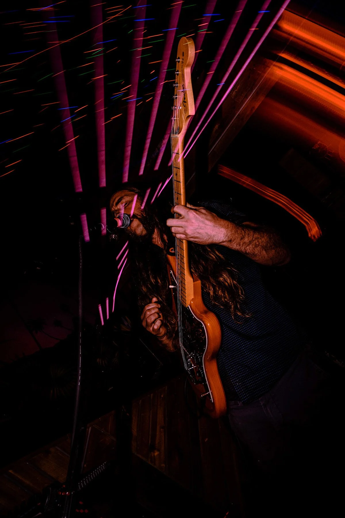 Chas Binns of Liquid Pennies singing into a microphone while playing an electric guitar with a wooden finish in a dimly lit setting with purple and orange light streaks.
