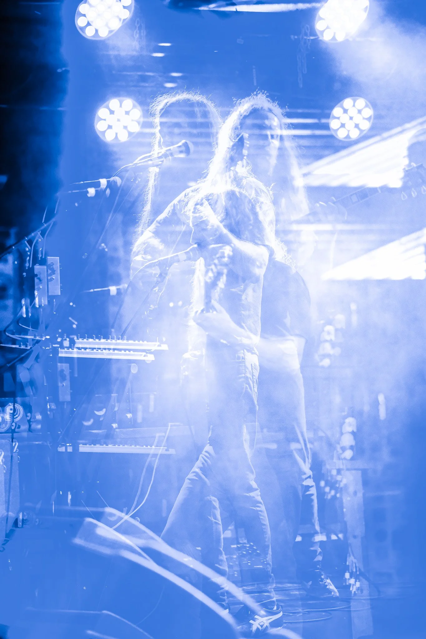 Two musicians with long hair performing on stage under blue lighting, with visible musical equipment and bright stage lights in the background.