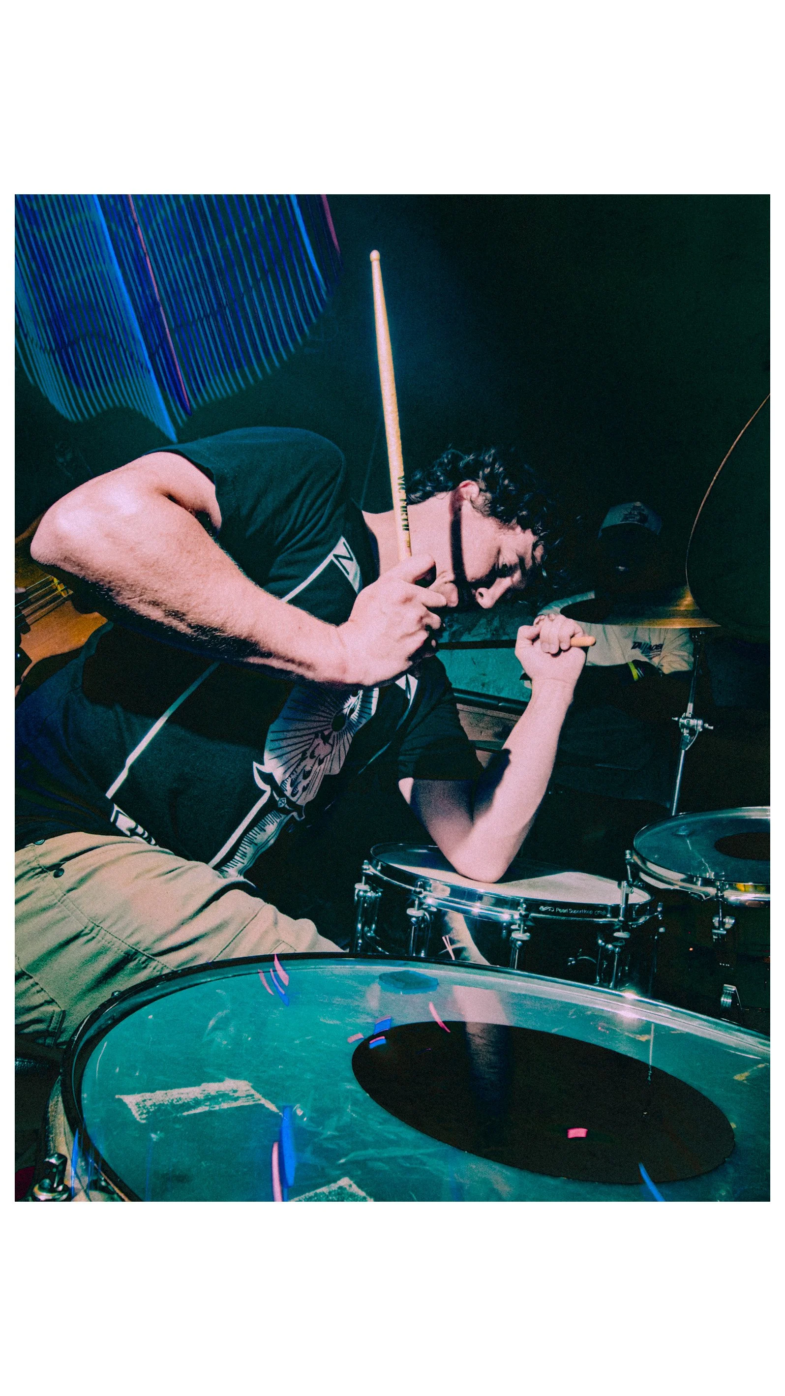 Pierson Whicker of Tampa Bay Psych Rock band Liquid Pennies playing drums passionately on stage, with a drum set in front of him and vibrant, colorful lights in the background.