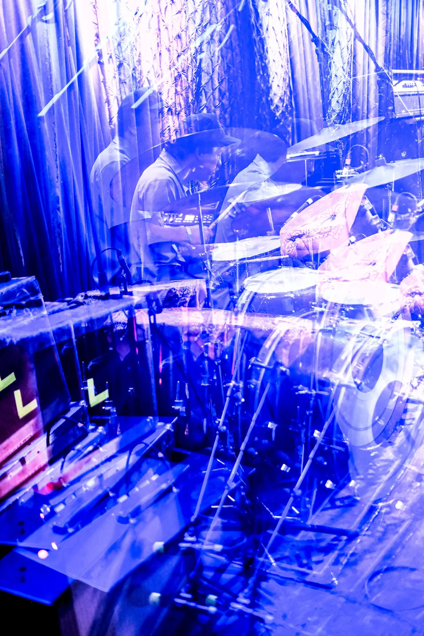 A blues and purple-tinted photo of a band performing on stage with multiple musicians, drums, and musical equipment visible through a reflective surface.