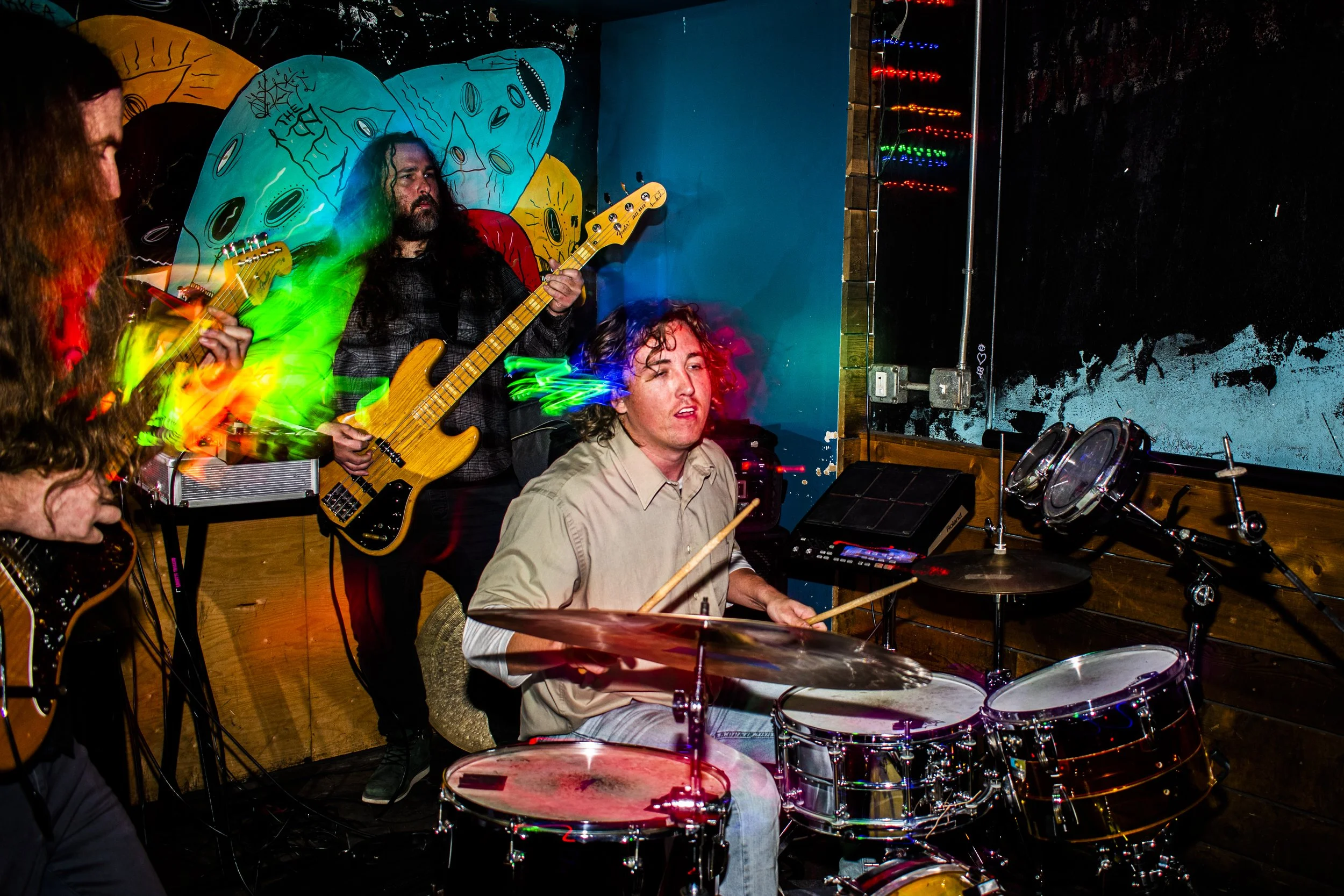 Rock trio Liqud Pennies performing live in a colorful, artistic venue. Pierson Whicker is playing drums, Tysonious Mink is playing bass guitar, and Chas Binns is playing an electric guitar. The background features colorful lighting effects.
