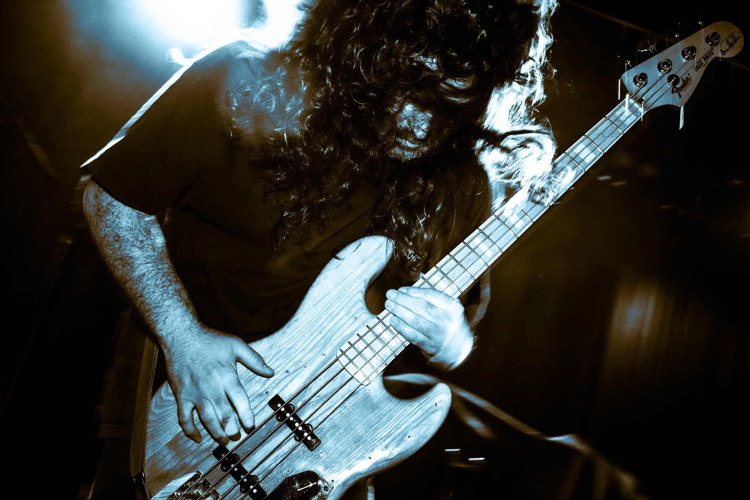 A person with long curly hair playing an electric bass guitar on stage, illuminated by blue lighting.