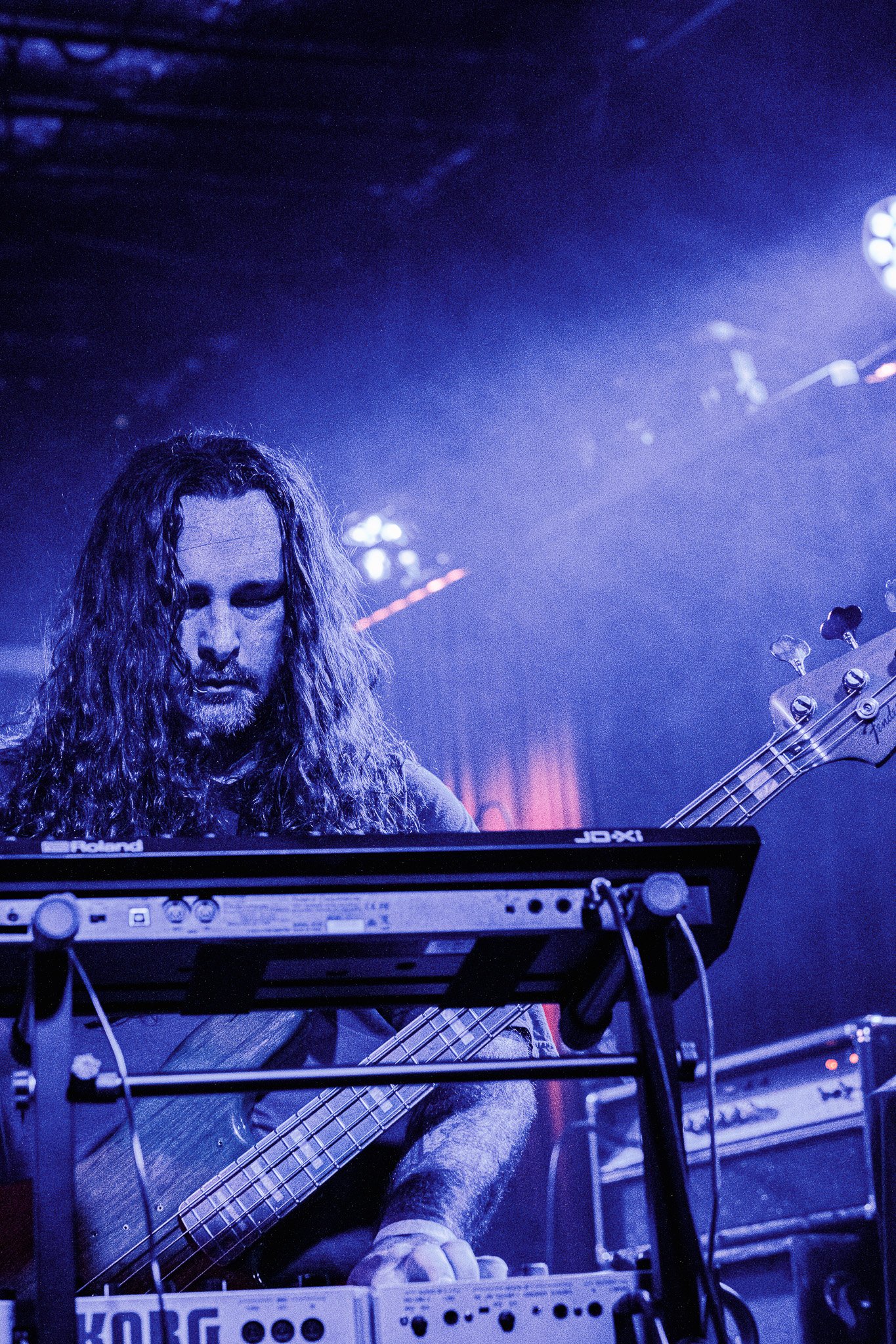 Tysonious Mink of the band Liquid Pennies with long curly hair playing a bass guitar on stage, with electronic equipment and stage lights around him.