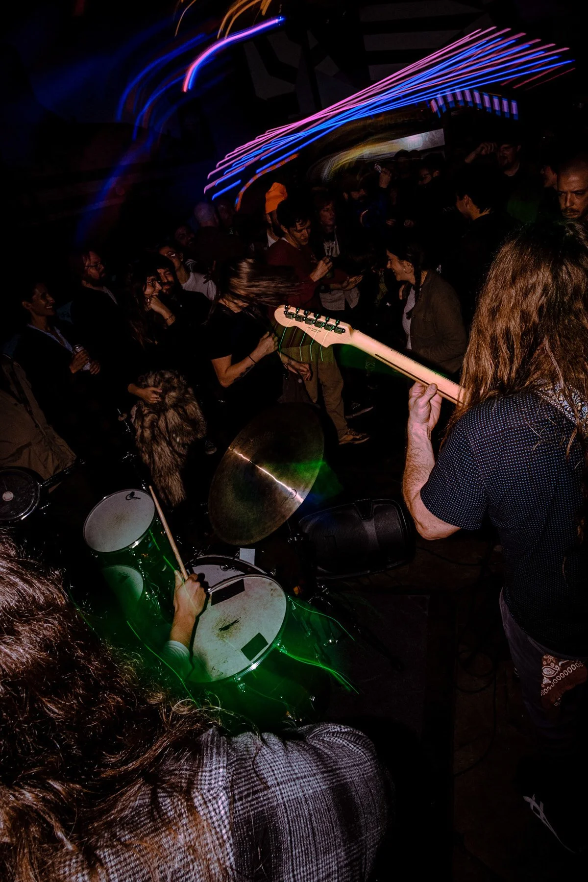A live music performance with a guitarist and drummer playing in front of a crowd at a dark venue, with colorful light streaks overhead.