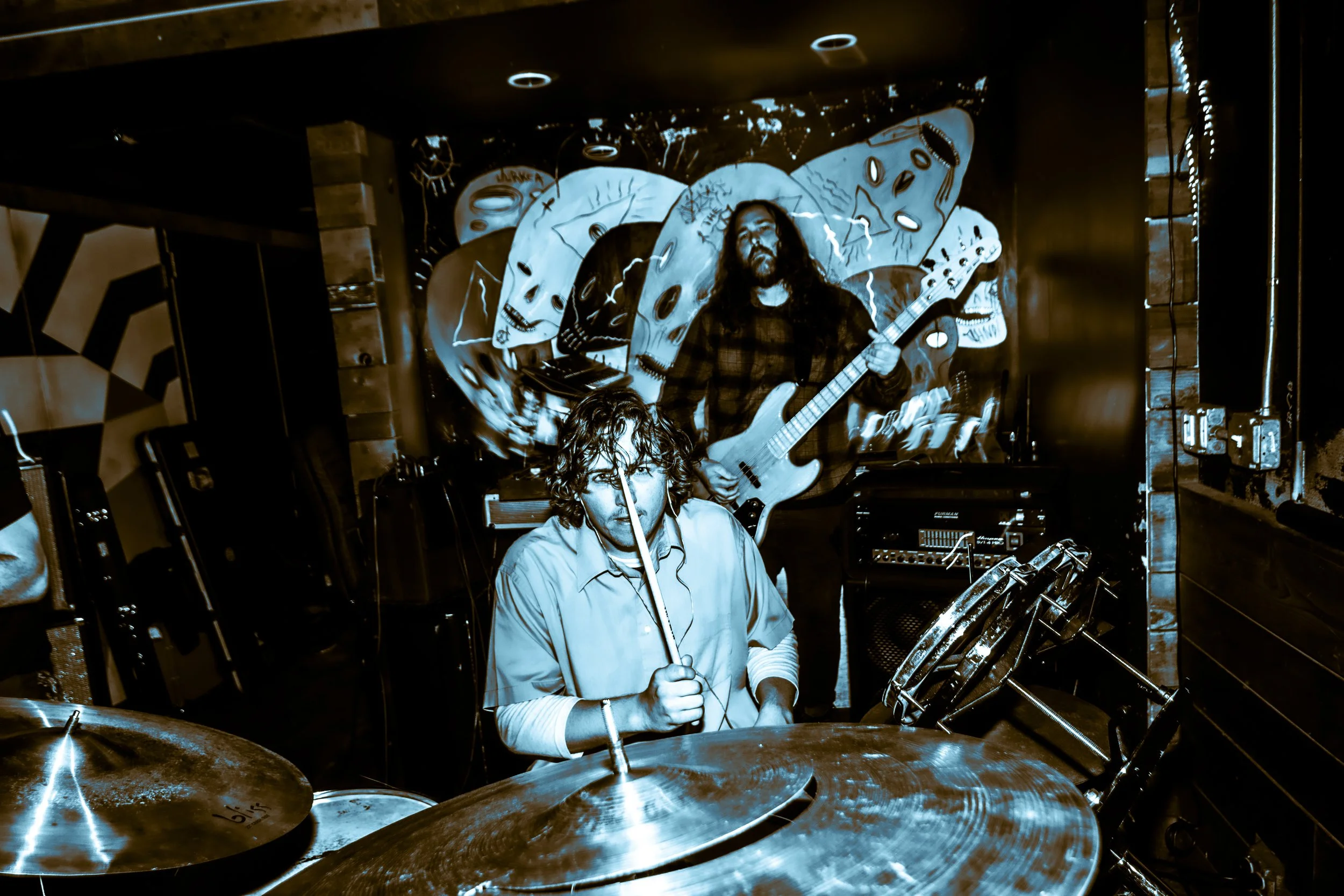 Two musicians performing on a stage with a colorful, skull-themed mural in the background. One is playing drums and the other is playing a bass guitar.