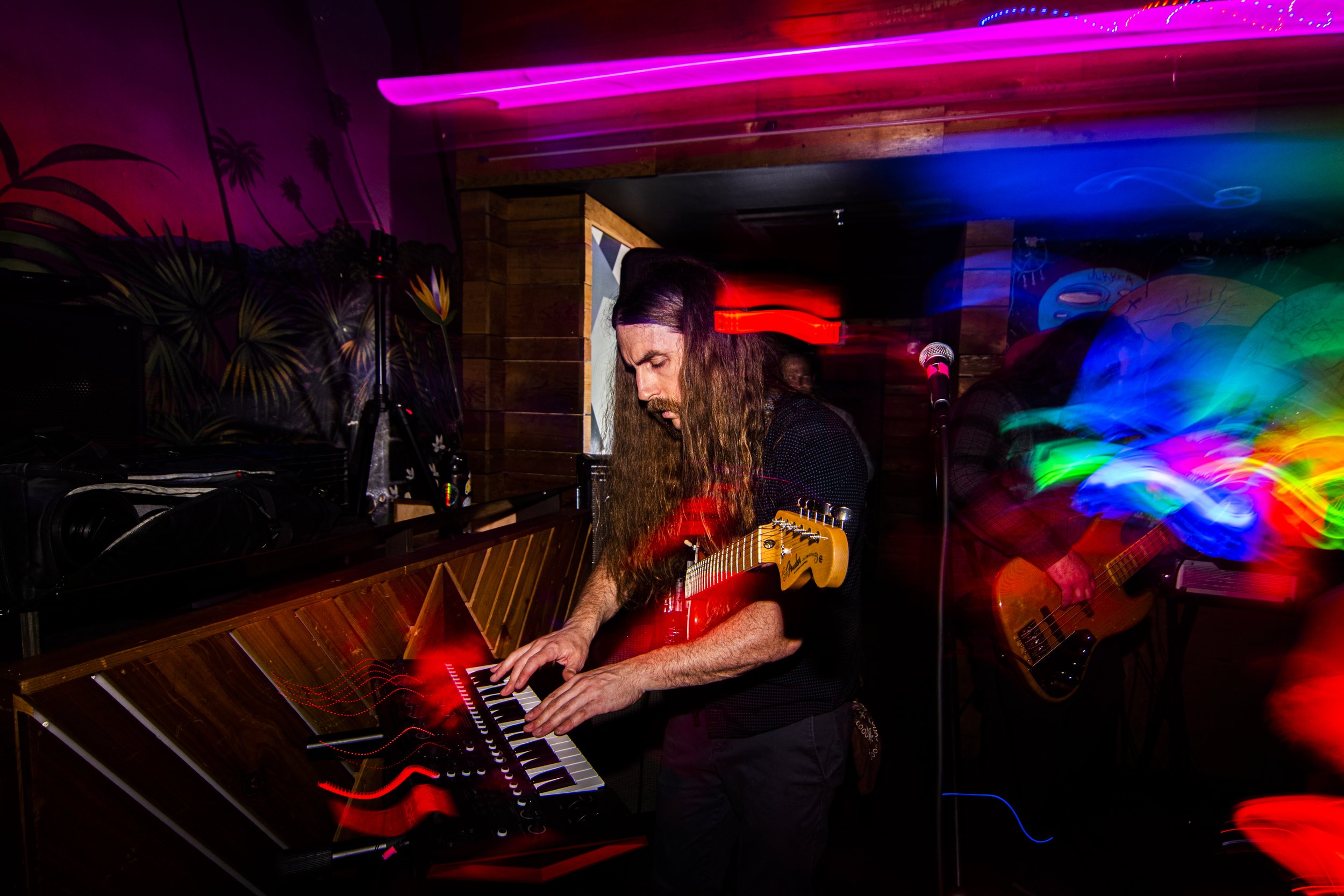 Musician with long hair playing synthesizer with colorful light streaks in a dimly lit room.