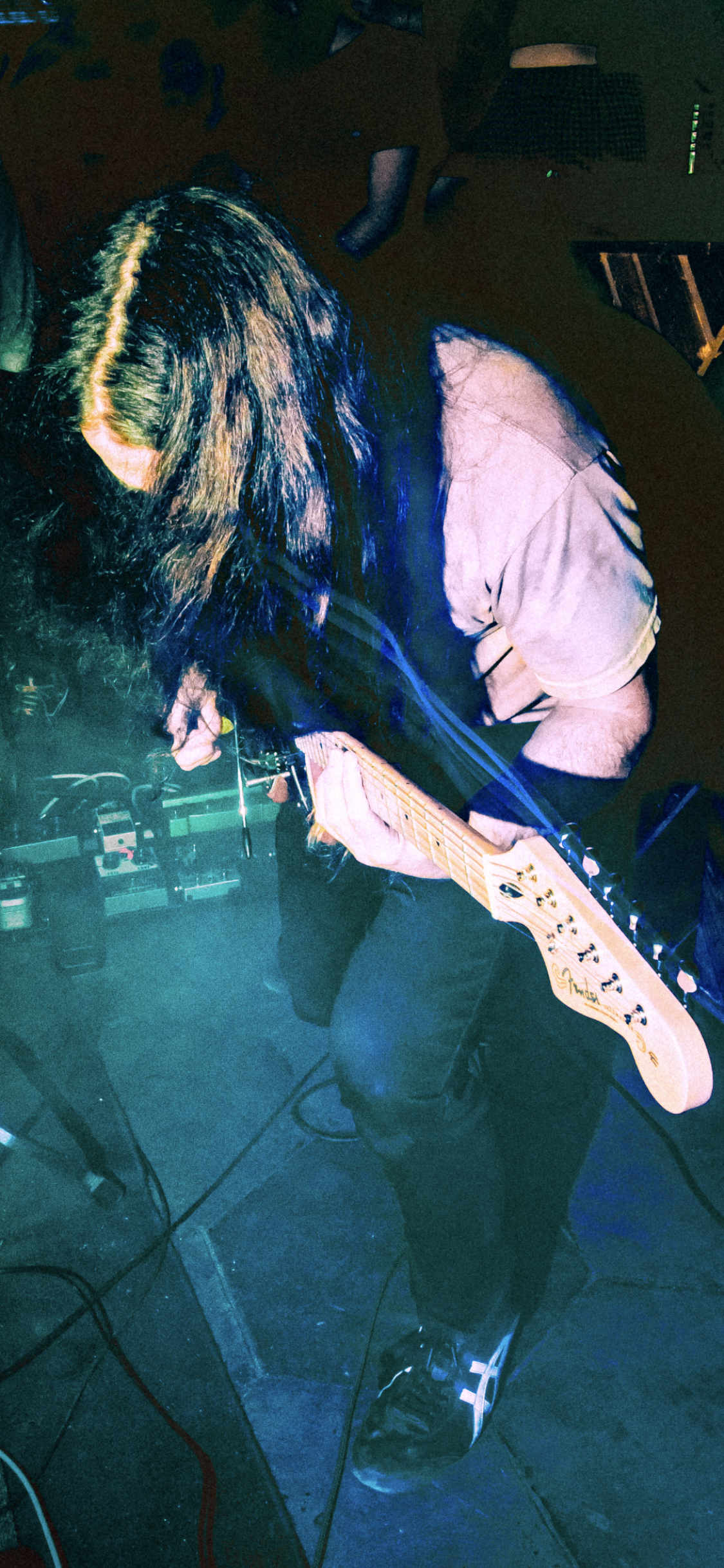 Musician playing electric guitar on stage, with dark background and stage equipment visible.