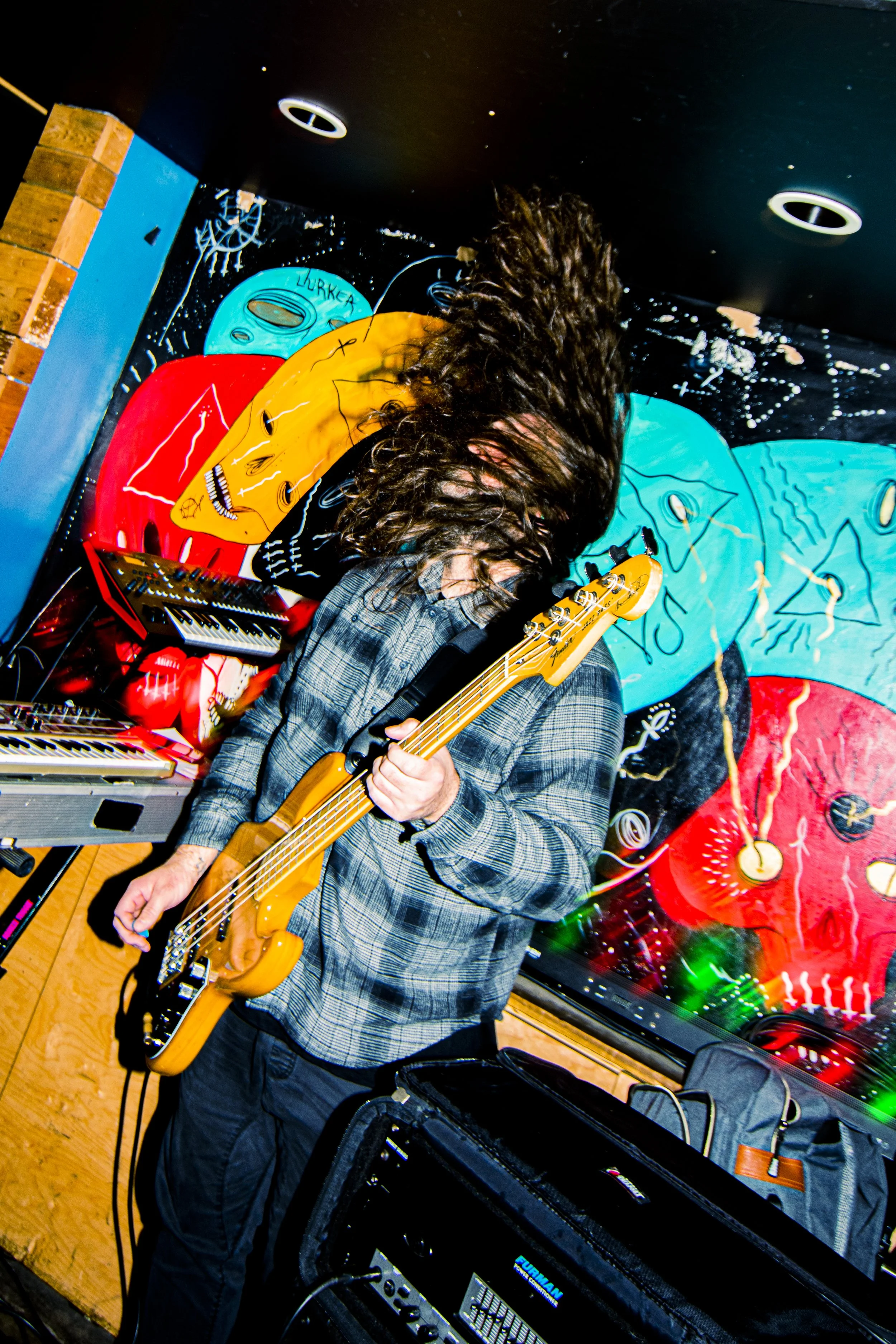 Musician with long curly hair playing an electric bass guitar on stage, surrounded by electronic equipment and colorful abstract wall art.