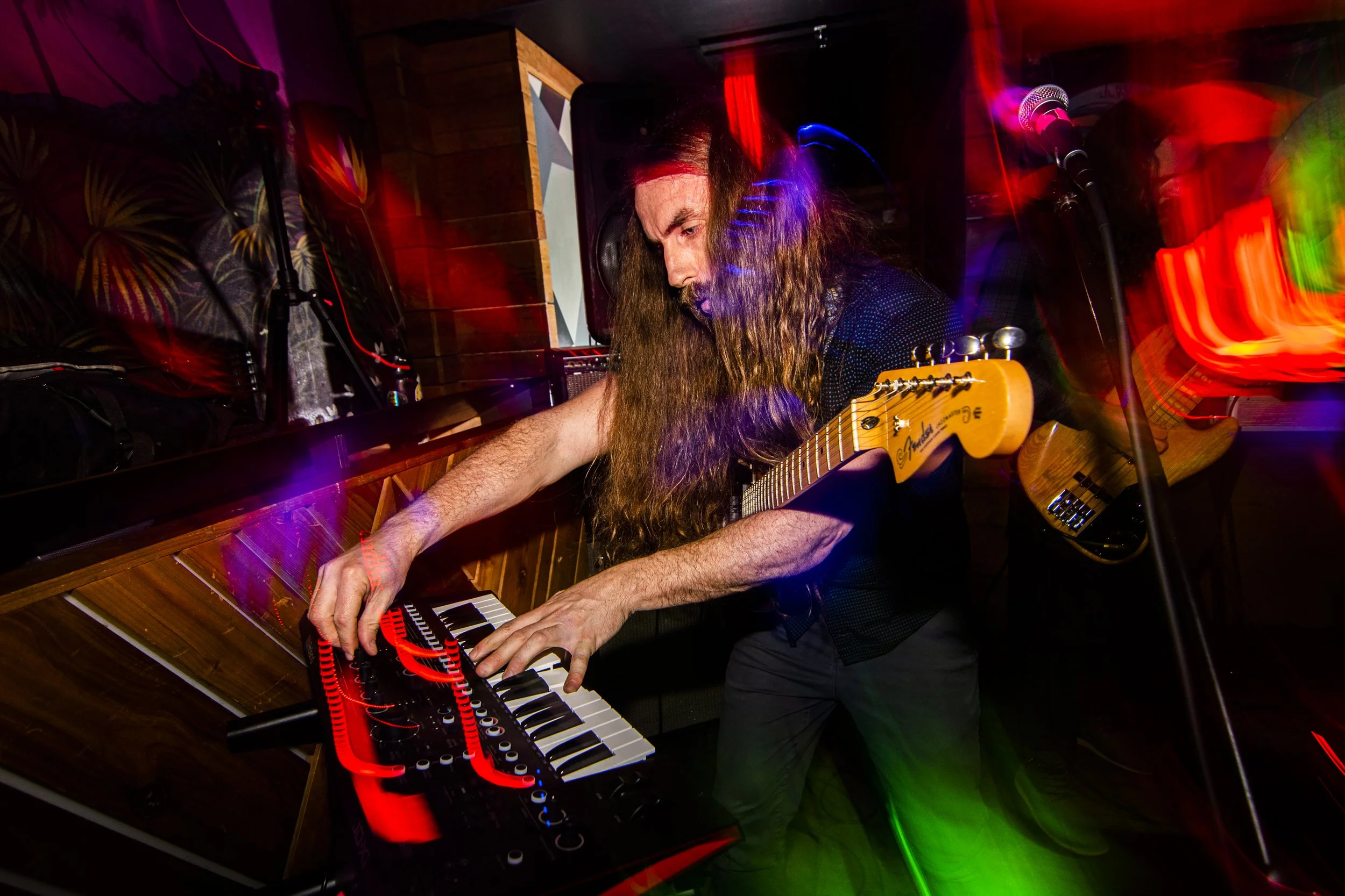 Musician with long hair playing keyboard and guitar in a dimly lit, colorful club with motion blur and light streaks.