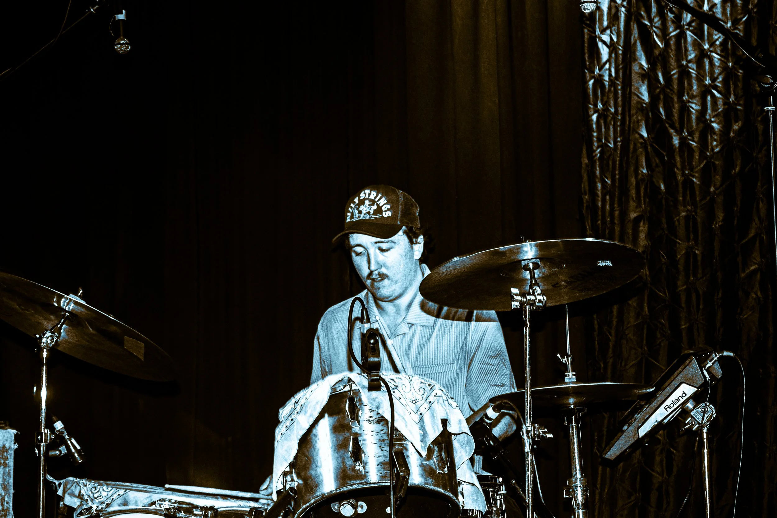 Psych rock band Liquid Pennies drummer PIerson Whicker playing drums, wearing a cap, on stage with dark curtains.