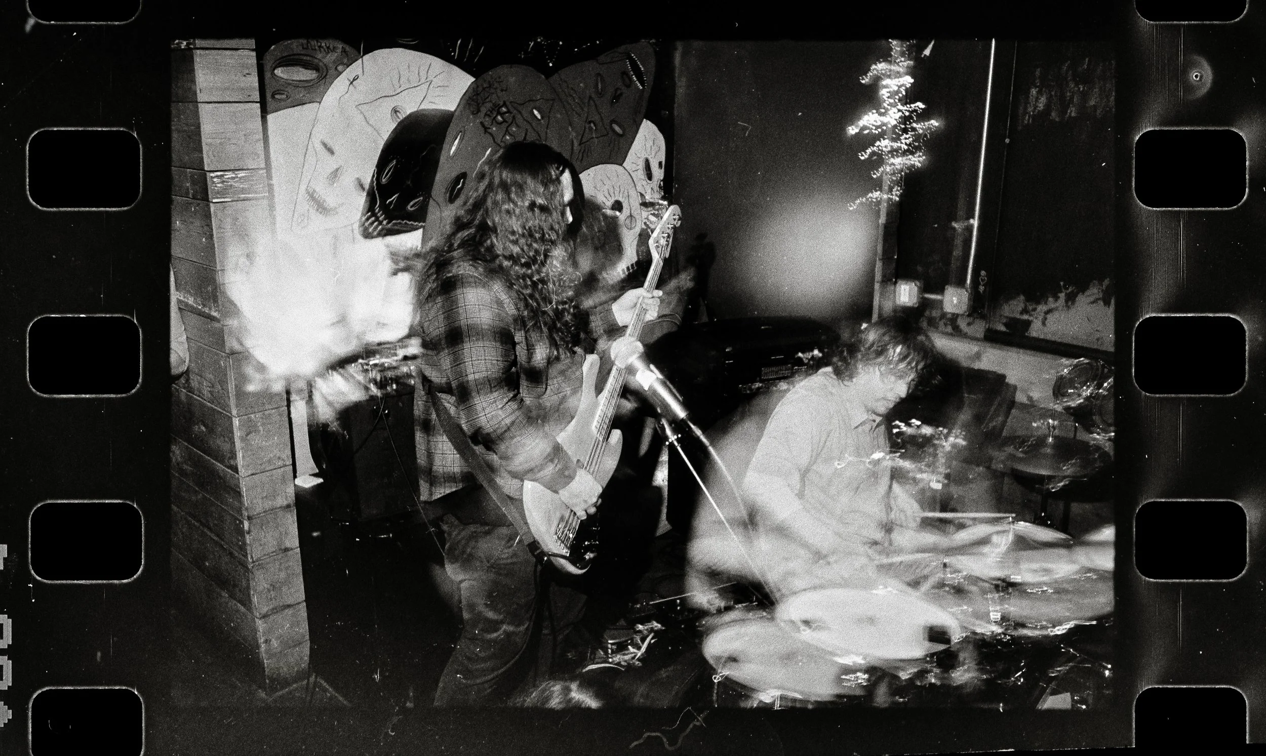 Black and white photo of a band performing in a small venue. A woman with long curly hair is playing an electric guitar and singing into a microphone. A man with long hair is seated at a drum set, and another person is partially visible on the left. 