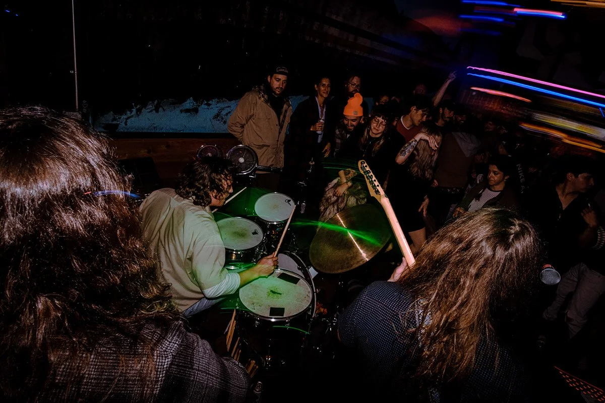 A drummer playing on stage at a crowded party or concert with colorful light streaks in the background.