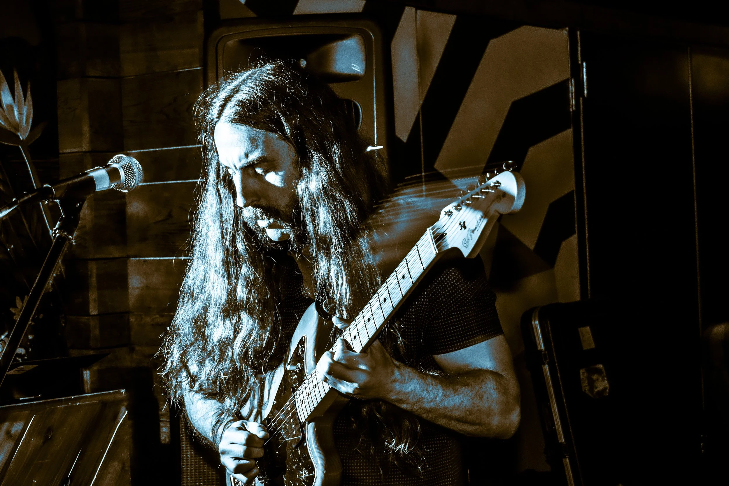 A man with long hair playing an electric guitar on stage, with a microphone in front of him and a speaker in the background.