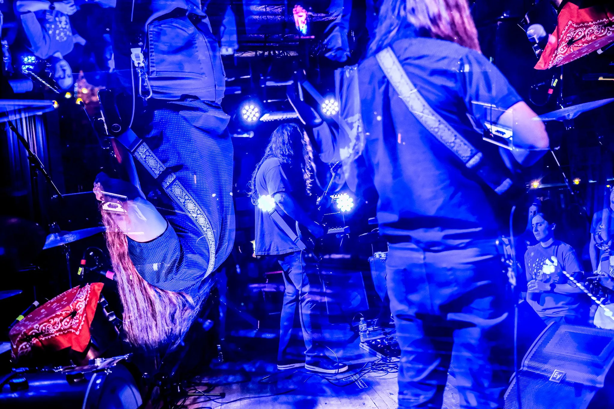 Musicians performing on stage with blue lighting, including a guitarist with long hair, and a nearby seated woman wearing a dark shirt, in a lively concert setting.