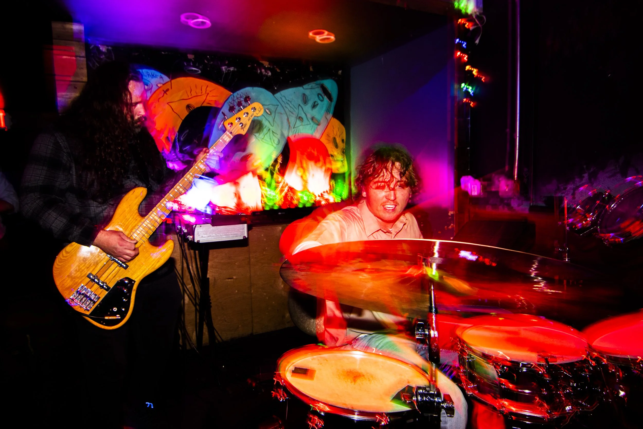 Two musicians performing live with vibrant, colorful lights and blurred motion; one playing a bass guitar and the other playing drums.