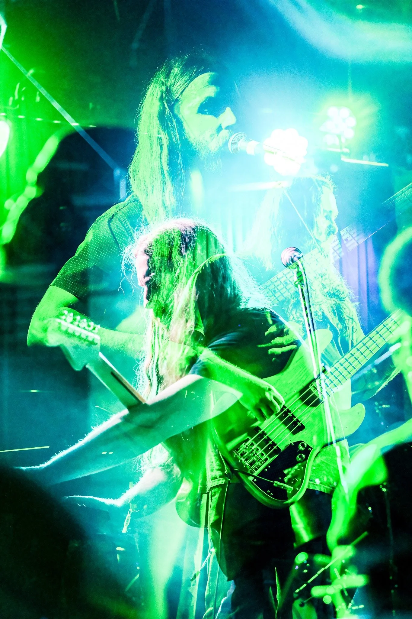 Two musicians performing on stage with vibrant green and blue lighting, one singing into a microphone and the other playing a bass guitar.