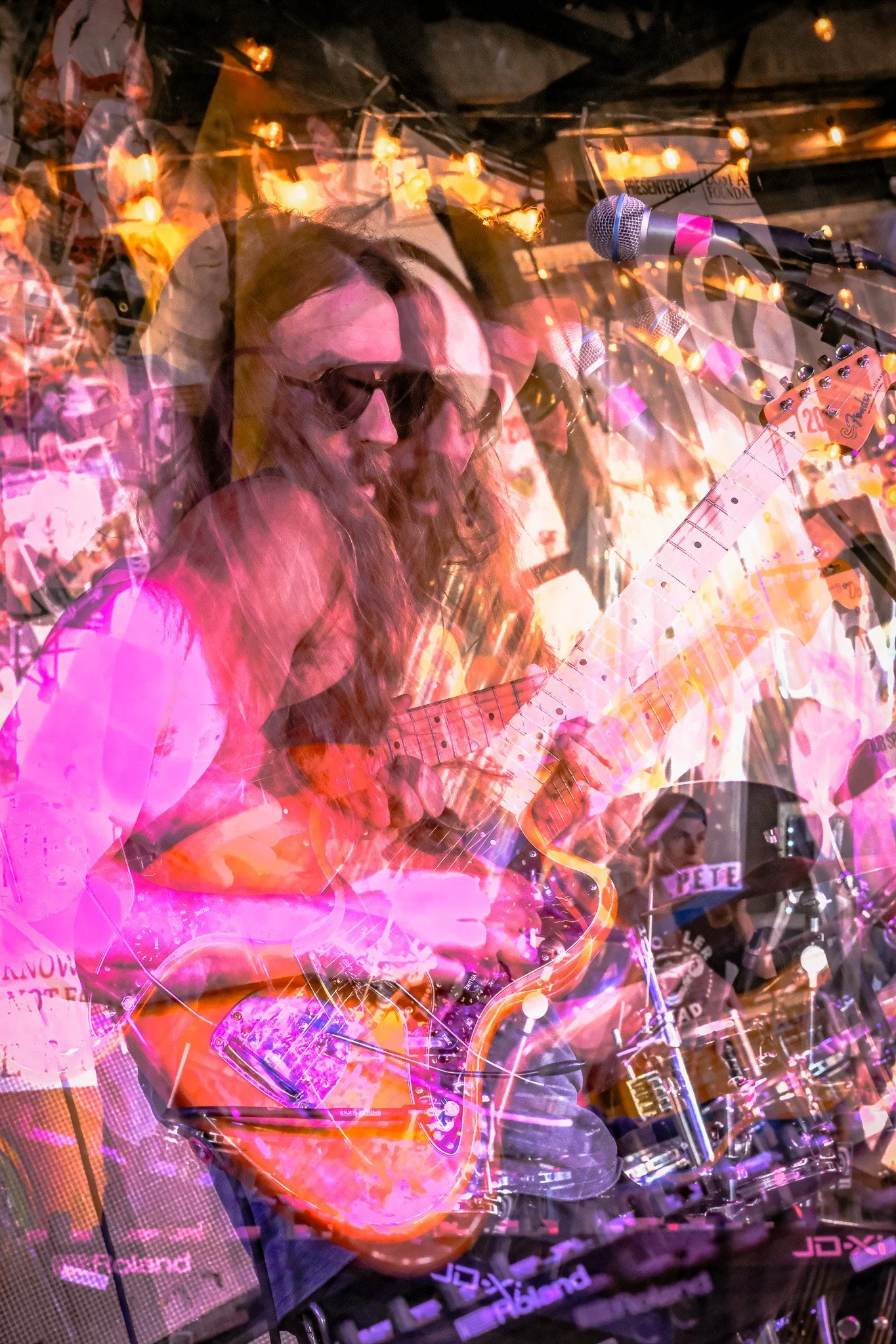 A musician with long hair and sunglasses is playing an electric guitar and singing into a microphone at a lively event with colorful lights.