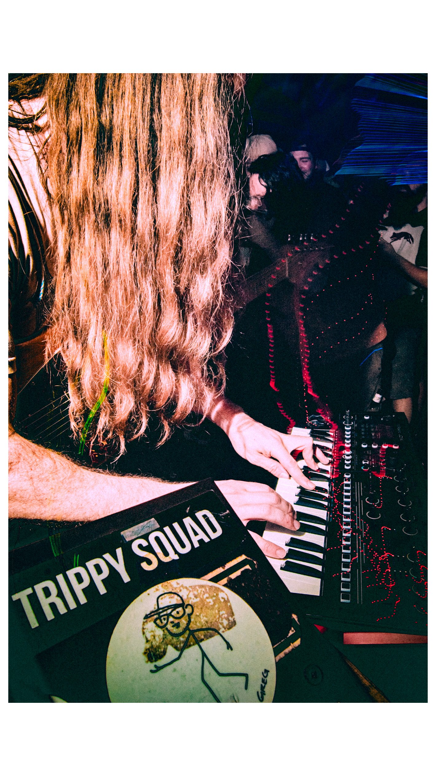 Chas Binns of the Neo-Psych rock band Liquid Pennies with long wavy hair playing a keyboard at a live concert with a sticker that reads 'TRIPPY SQUAD' and a drawing of a person with sunglasses and curly hair.