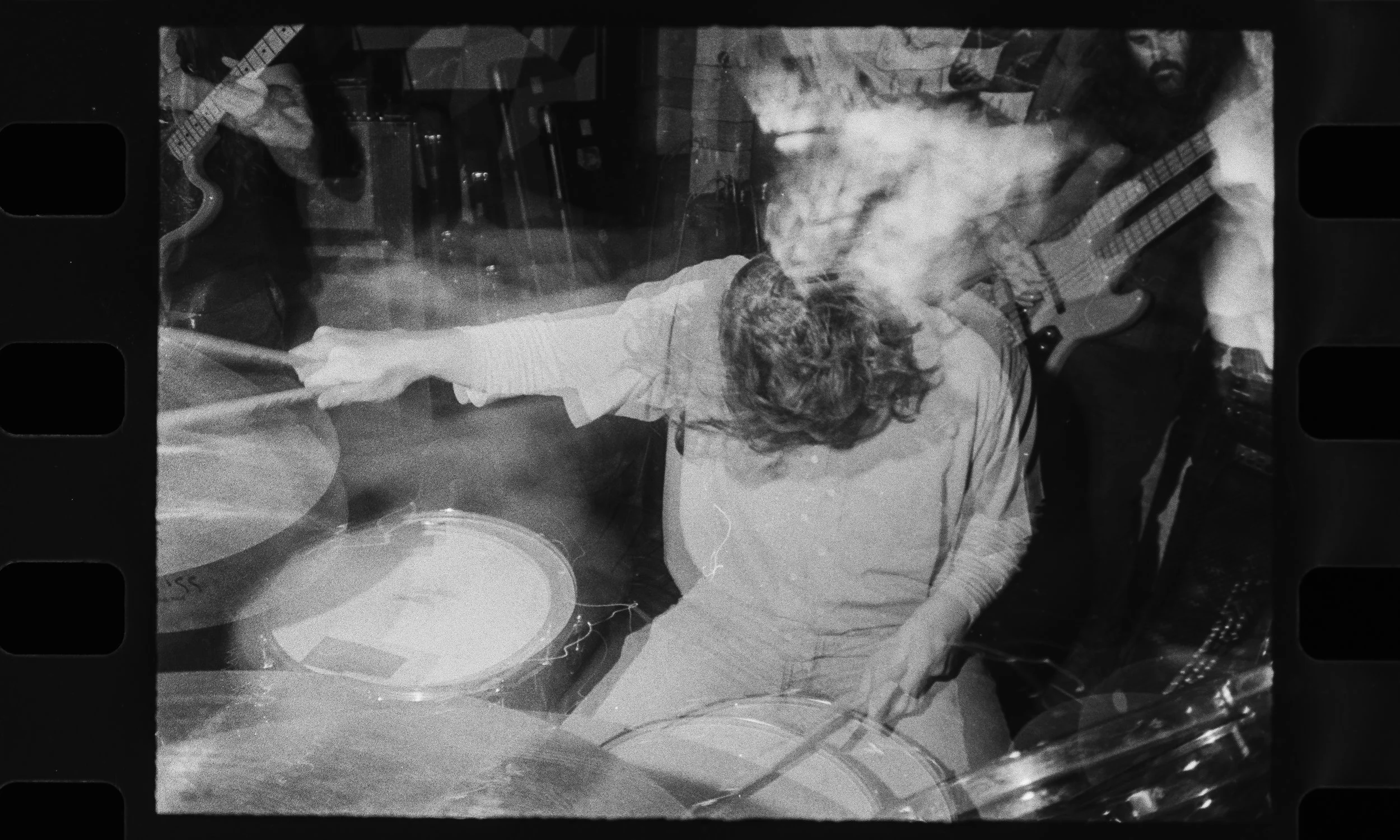 Black and white photo of a drummer and two guitar players performing on stage. The drummer is in the center, with one guitarist on each side, all engaged in playing their instruments.