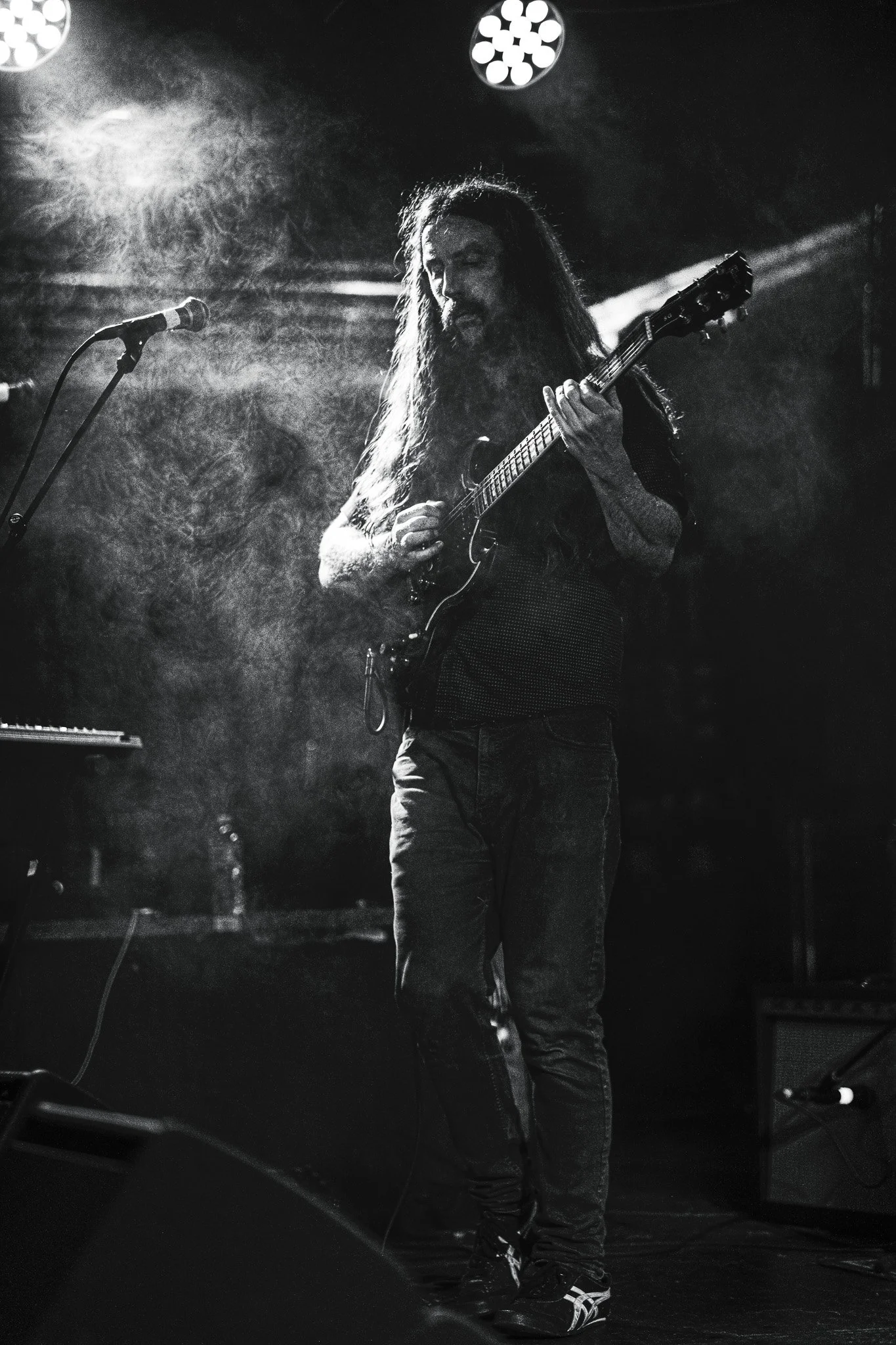 Black and white photo of a long-haired man playing an electric guitar on stage. There is a microphone stand in front of him and stage lights overhead.