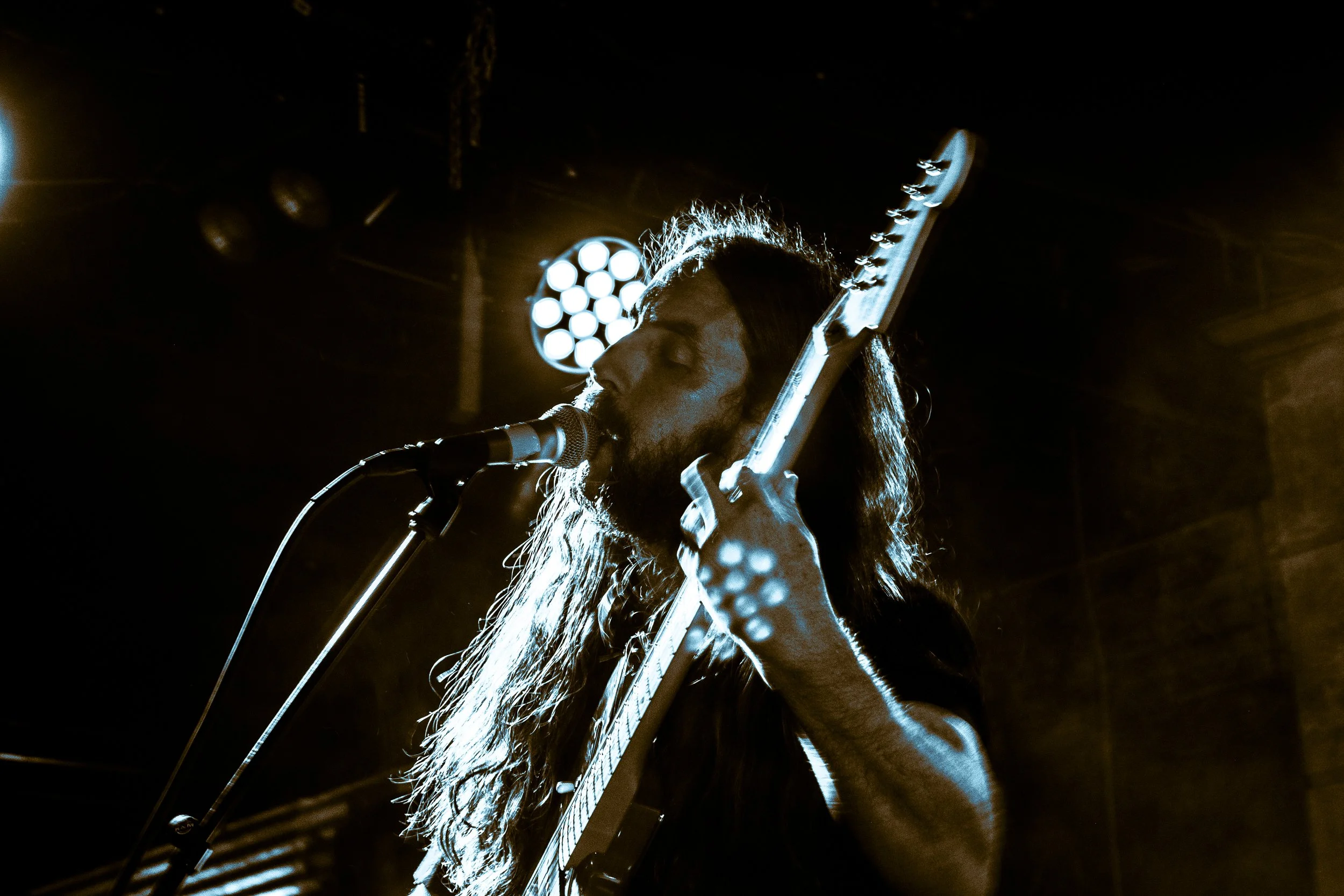 Chas Binns of the band Liquid Pennies singing into a microphone, playing electric guitar on stage with bright stage lights in background. From the Liquid Pennies official website.