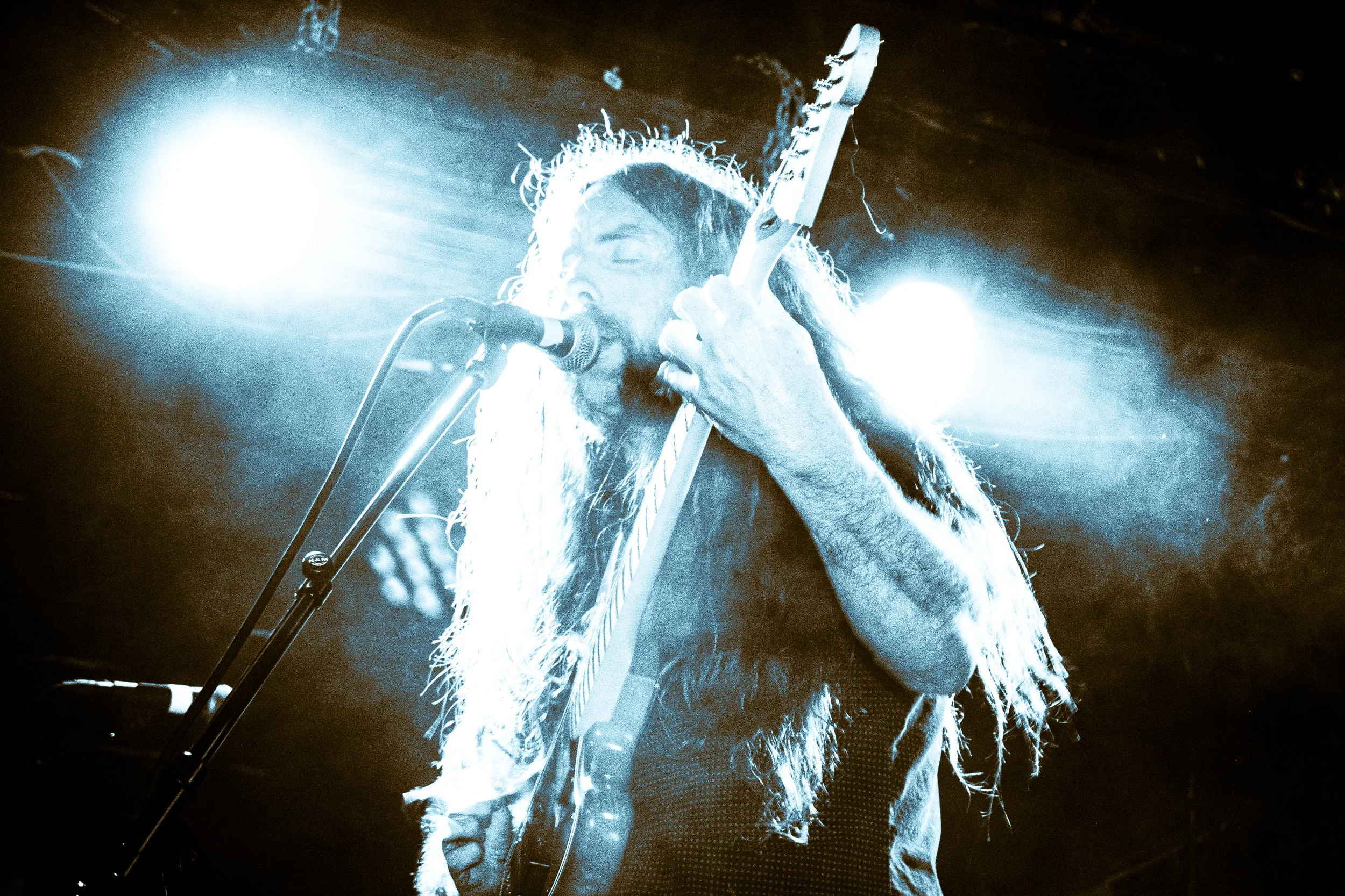 Rock band Liquid Pennies frontman/guitarist Chas BInns playing an electric guitar and singing into a microphone on stage, illuminated by bright stage lights.