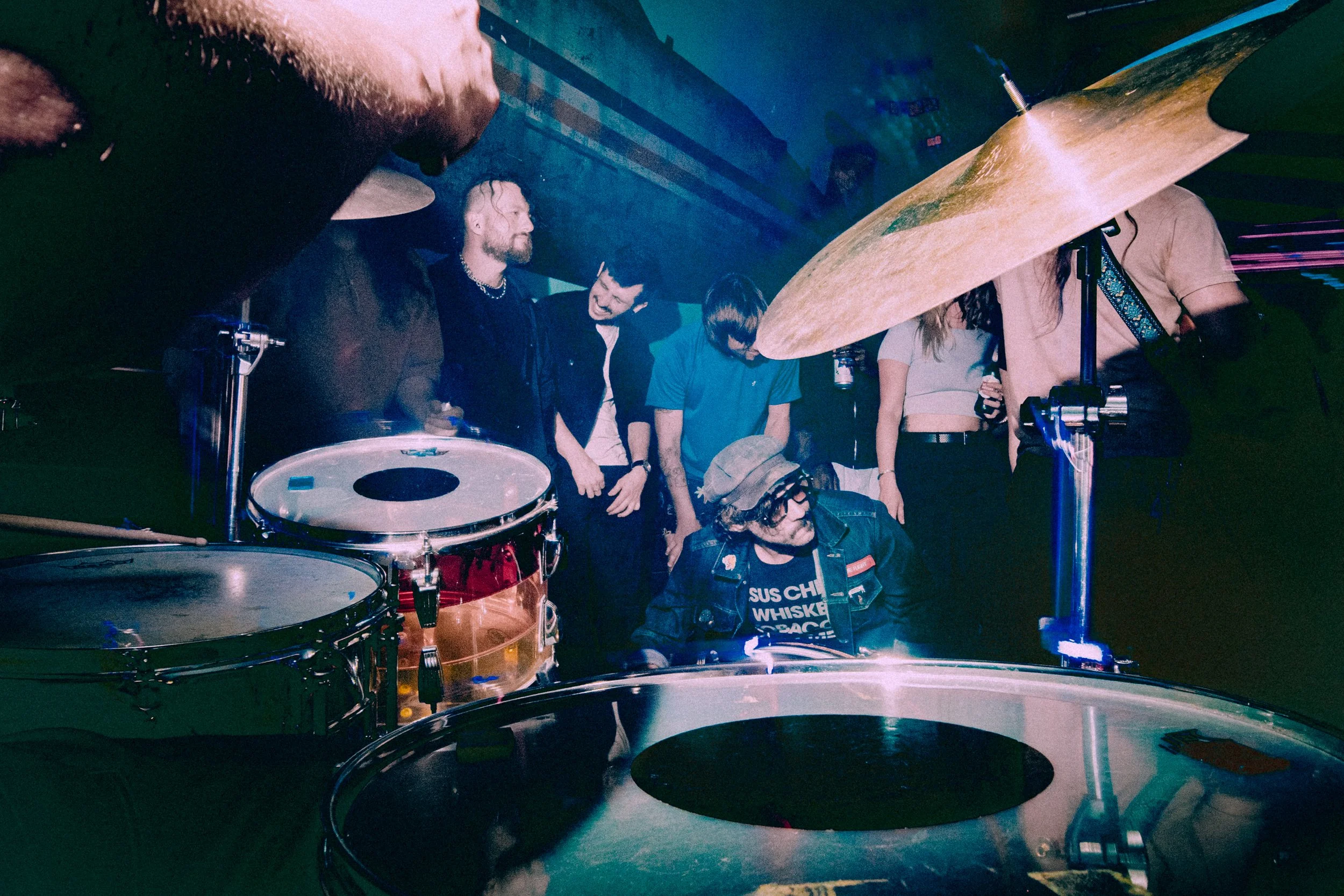 A drummer wearing glasses and a hat is playing a drum set at a party or concert, surrounded by people dancing and socializing in a dimly lit venue with blue and purple lighting.